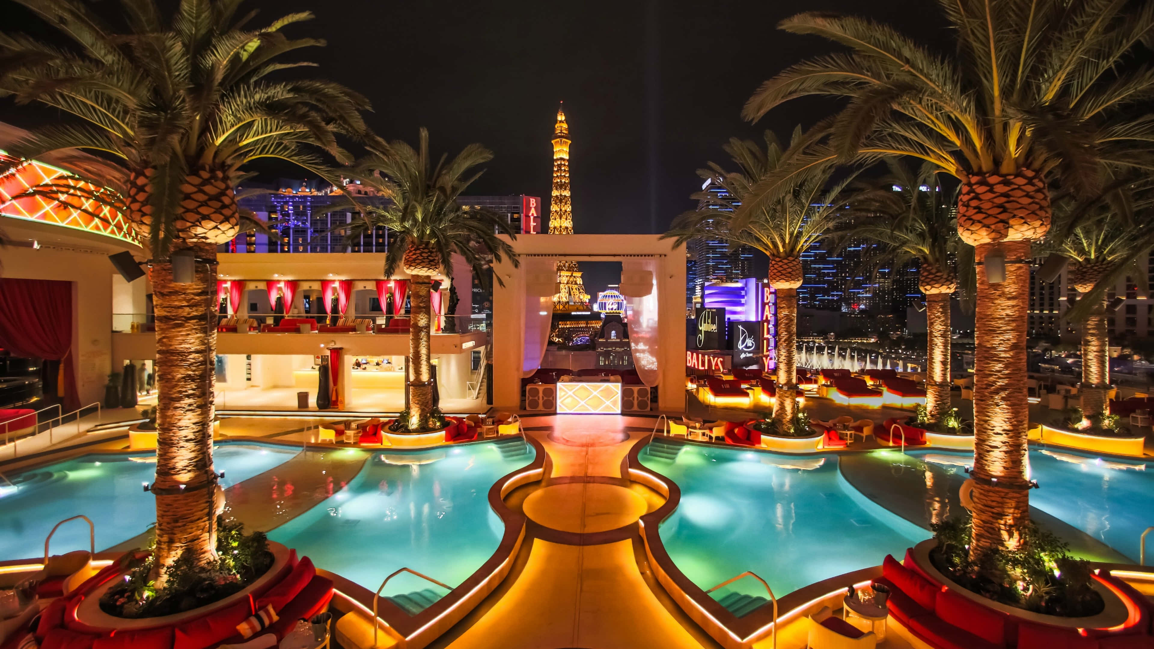 4K Las Vegas 桌布 壁紙 — 一個迷人的夜間度假村泳池區，由溫暖的燈光照亮，棕櫚樹和複製的艾菲爾鐵塔映襯著閃爍的城市天際線。