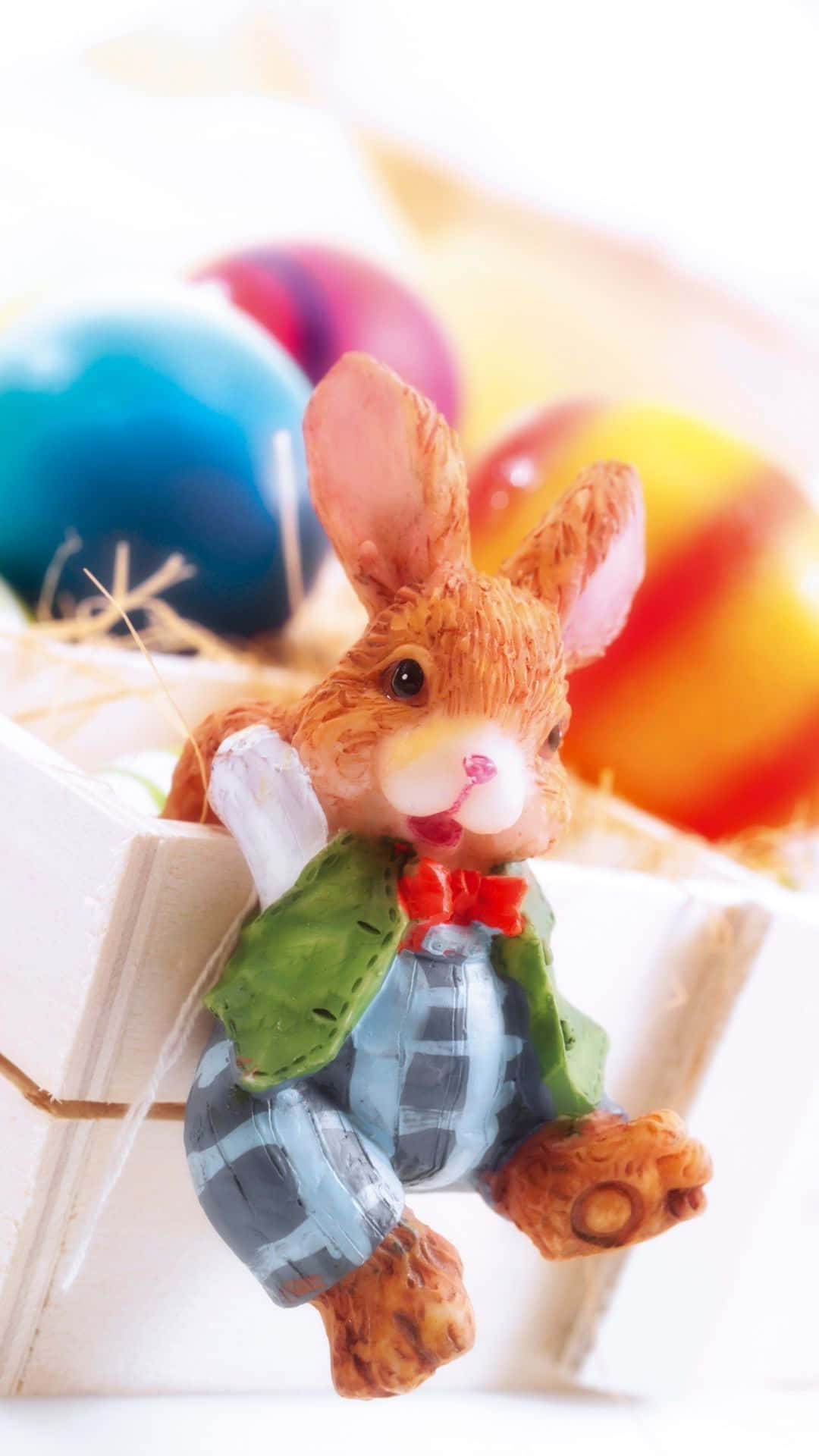 Easter Bunny 桌布 壁紙 — 一個迷人的棕色兔子公仔，穿著格子連身褲和綠色背心，坐在一個裝有彩色復活節蛋的木籃子裡。