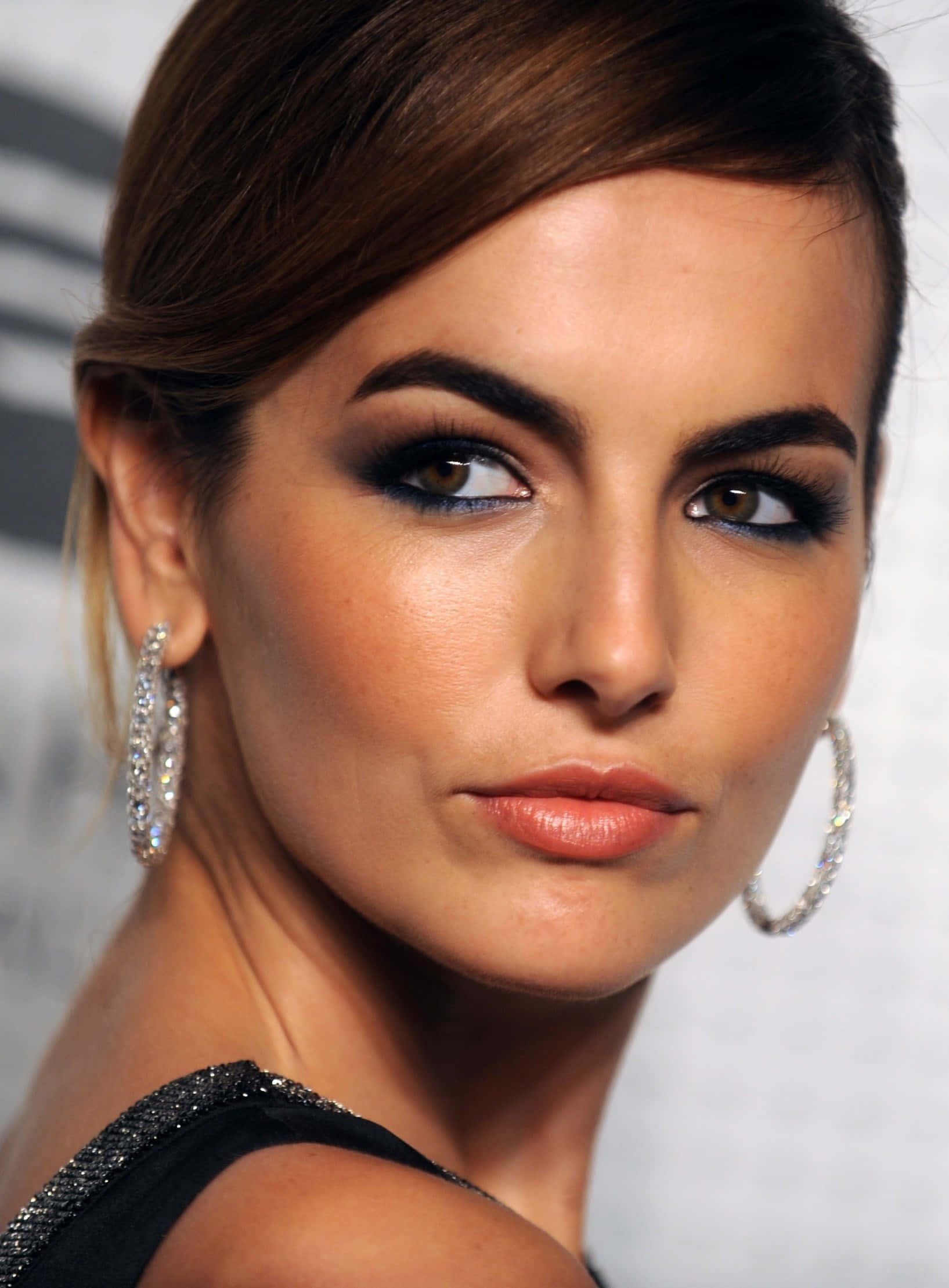Camilla Belle 桌布 壁紙 — 一個迷人的特寫鏡頭，拍攝一位化著大膽妝容、戴著閃亮圈圈耳環的女性，背景模糊。