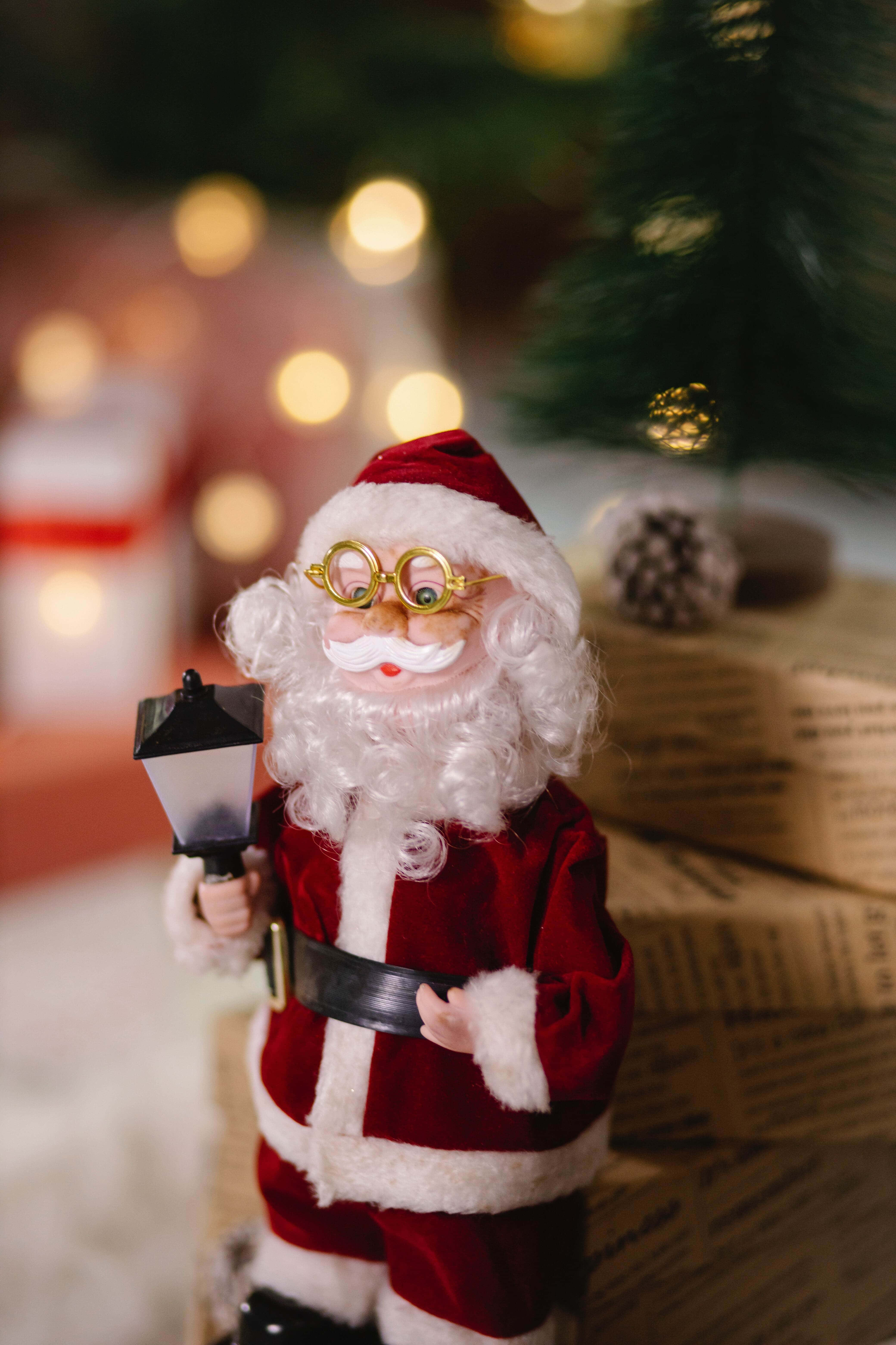 Santa Claus 桌布 壁紙 — 一個迷人的聖誕老公公公仔，戴著金邊眼鏡和拿著燈籠，站在裝飾好的樹和禮物旁邊。