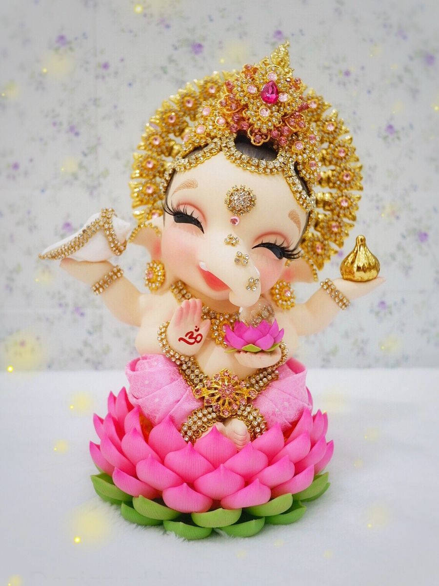 laxmi ganesh saraswati 桌布 壁紙 — 一個迷人裝飾的嬰兒象神雕像坐在粉紅色蓮花上，戴著黃金首飾和寶石皇冠，背景是花卉圖案。