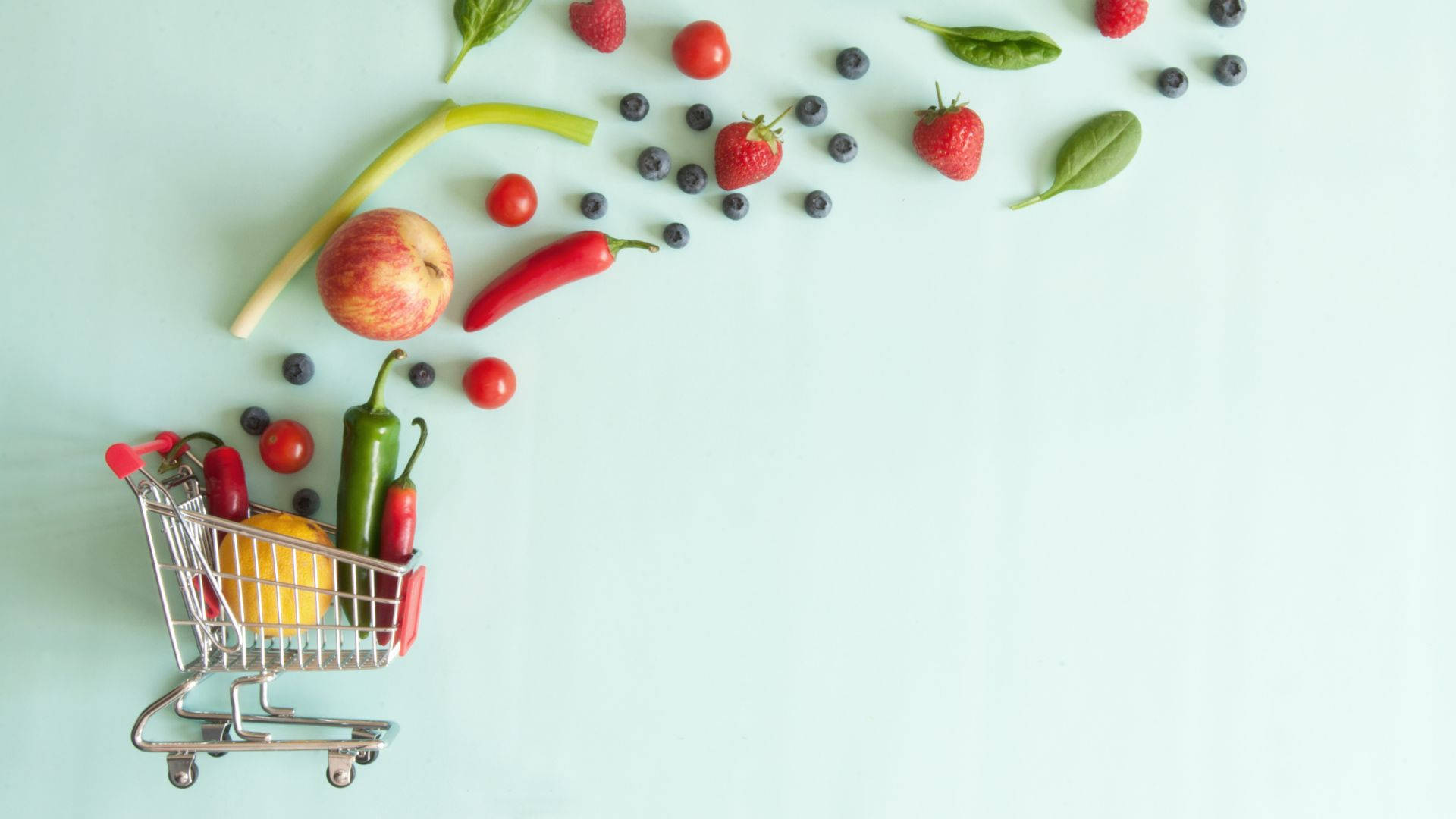 Fruits And Vegetables 桌布 壁紙 — 一個迷你購物車裝滿了色彩繽紛的新鮮農產品，在淡薄荷色背景上灑滿了鮮豔的水果和蔬菜。