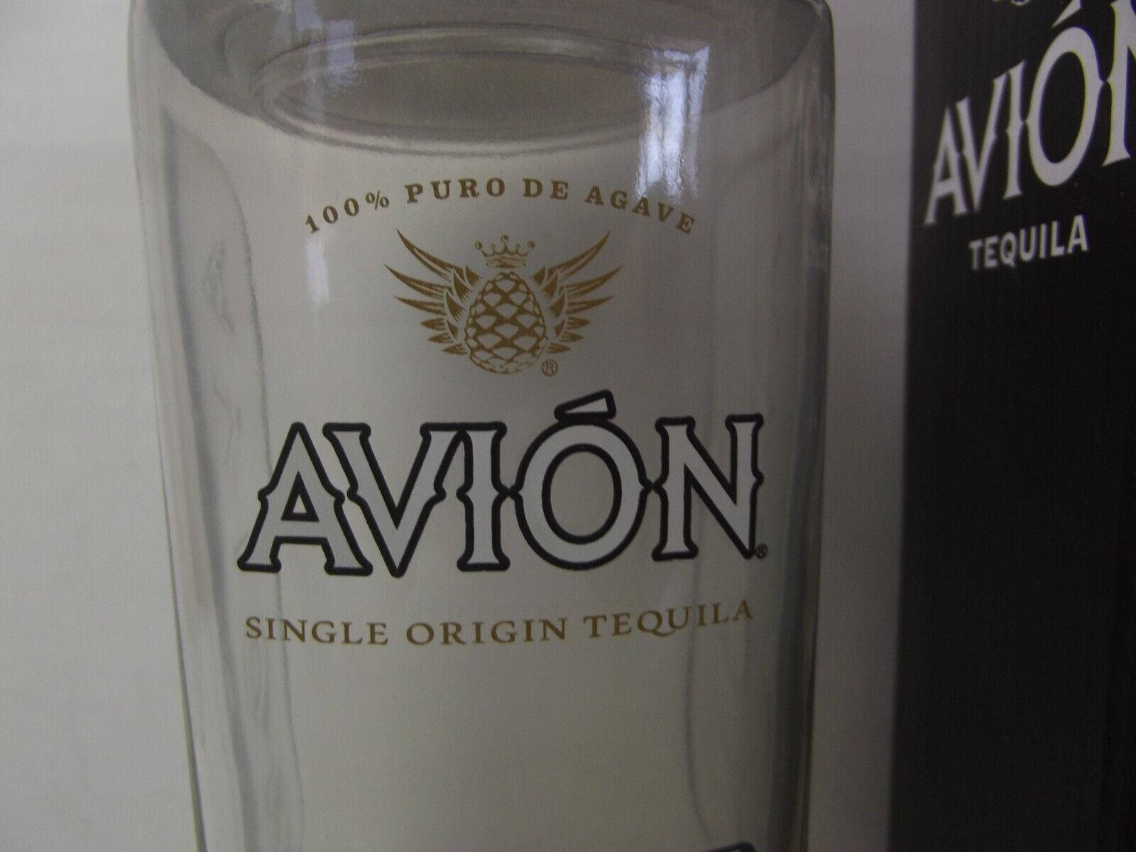 bottle 桌布 壁紙 — 一個透明Avión龍舌蘭酒杯的特寫，帶有金色和黑色品牌標識，旁邊是其黑色酒瓶。