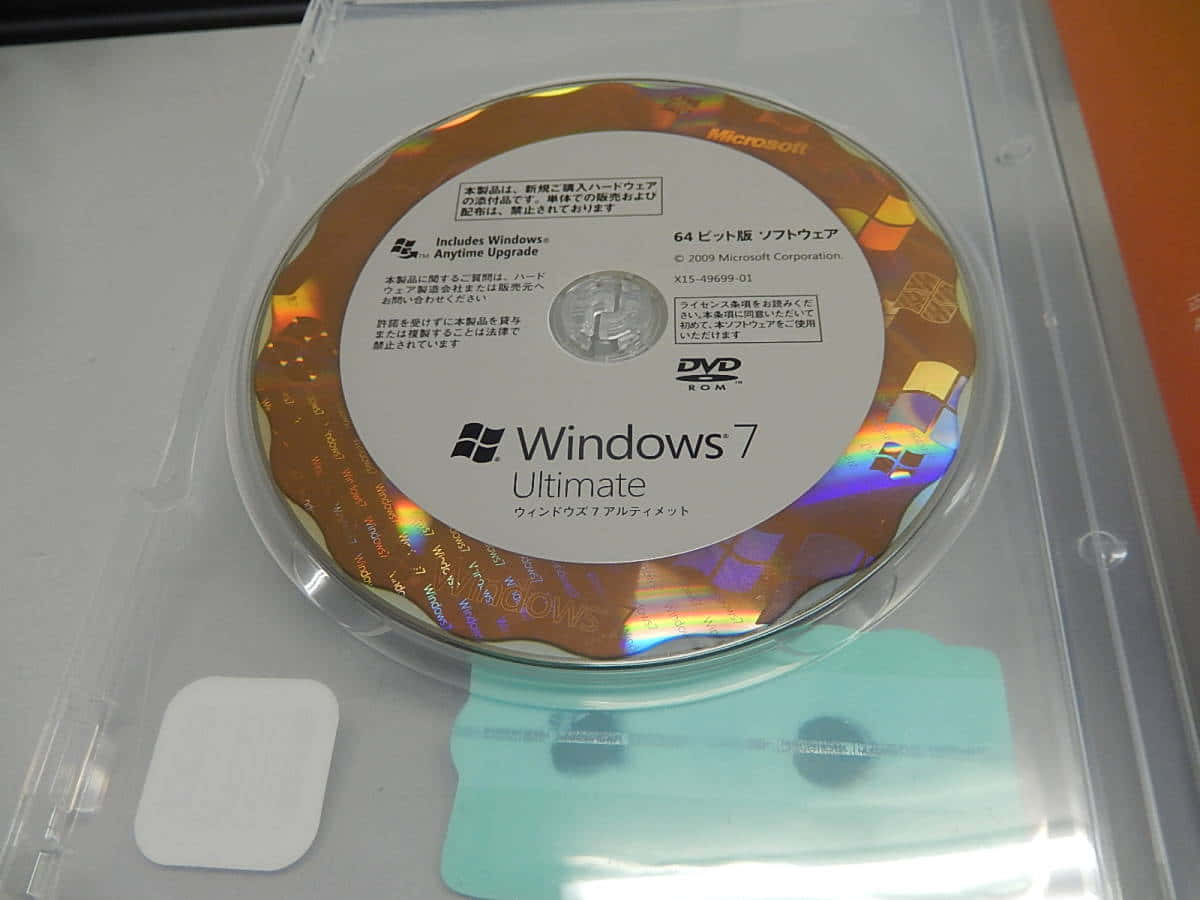 Windows 7 桌布 壁紙 — 一個透明塑膠盒裝的Windows 7 Ultimate DVD，上面有日文字和光碟的彩虹反射。