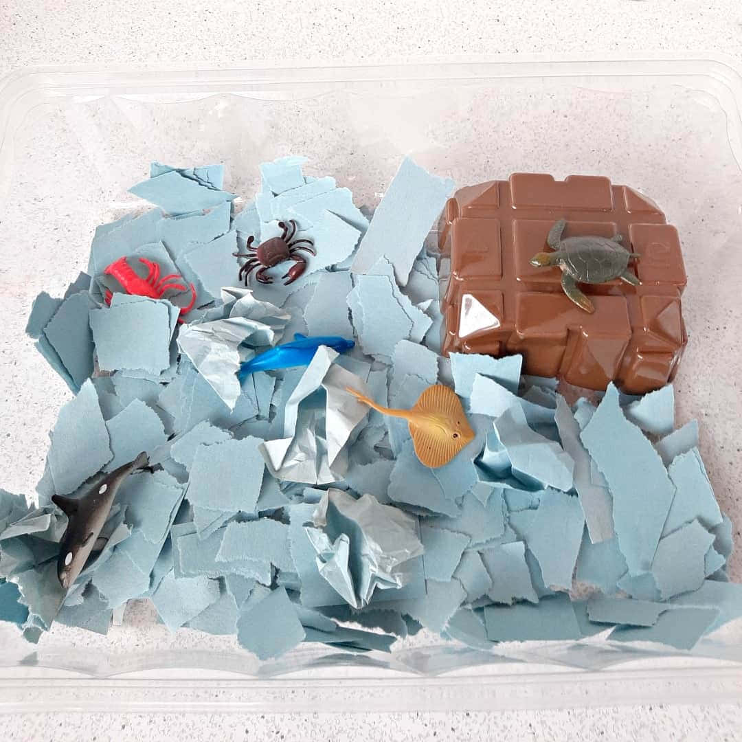 Torn Paper 桌布 壁紙 — 一個透明容器裝有藍色碎紙，內有塑膠海洋生物和一個棕色岩石狀結構。