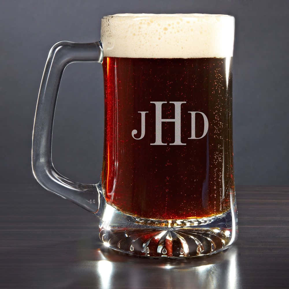 Beer 桌布 壁紙 — 一個透明玻璃啤酒杯，刻有JHD縮寫，盛載深琥珀色啤酒，頂部有白色泡沫，背景為深灰色。