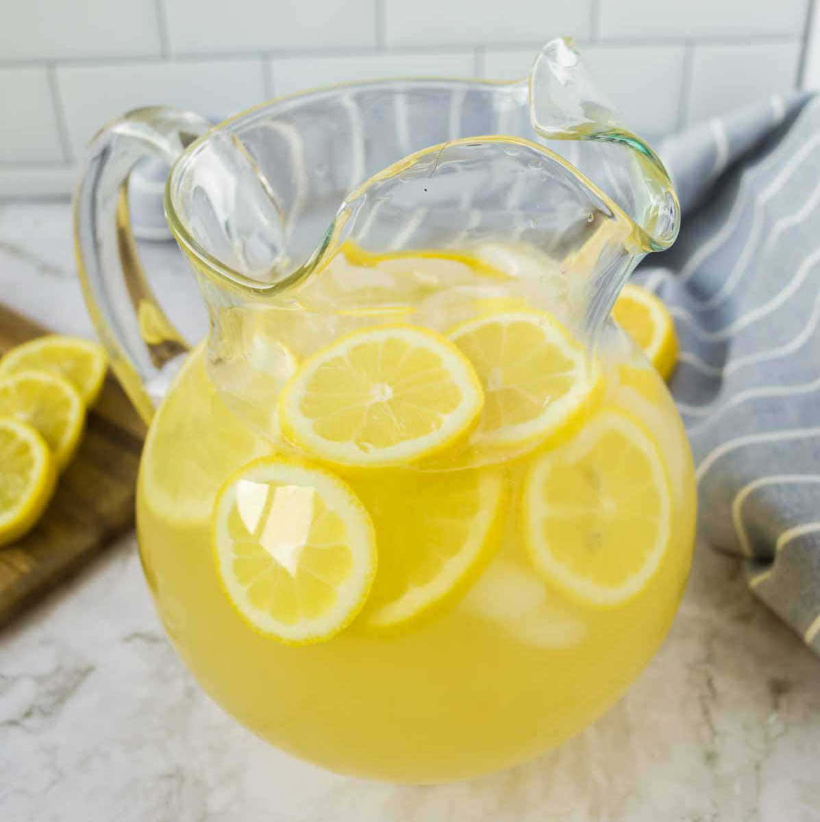Lemonade 桌布 壁紙 — 一個透明玻璃壺裝滿了鮮黃色的檸檬水和漂浮的檸檬片，放在大理石檯面上，旁邊有一條條紋毛巾和切好的檸檬。