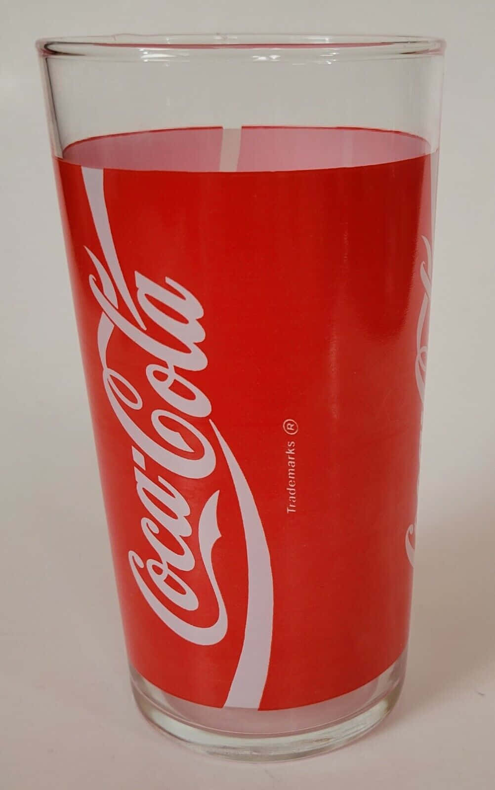 coca cola 桌布 壁紙 — 一個透明玻璃杯，帶有標誌性的紅色可口可樂品牌和白色字體標誌，背景為純白色。