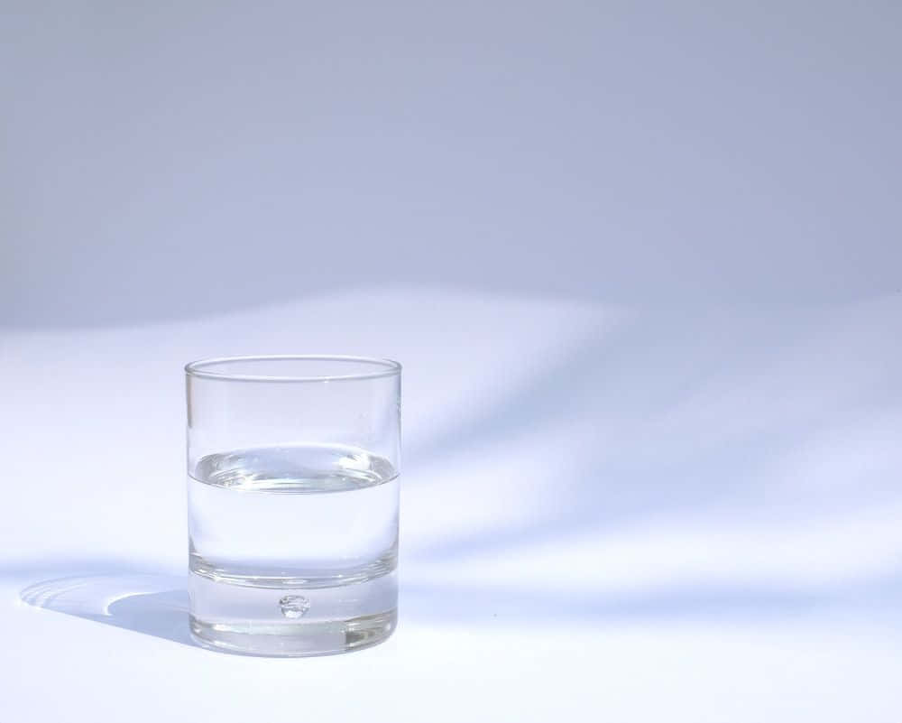 Glass Of Water 桌布 壁紙 — 一個透明玻璃杯半滿水，放在白色表面上，在明亮光線下投射出柔和陰影。