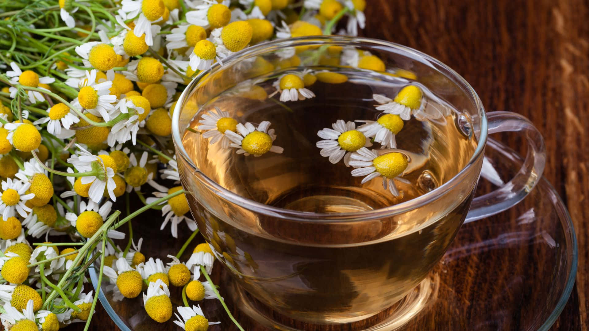 Floral Tea 桌布 壁紙 — 一個透明玻璃杯盛載著金黃色的洋甘菊茶，茶中有浮花，旁邊木桌上放著一束鮮花。