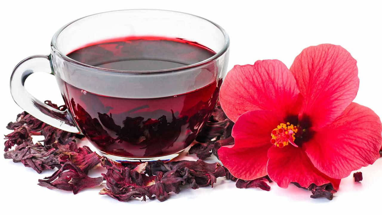 Hibiscus 桌布 壁紙 — 一個透明玻璃杯裝著深紅色洛神花茶，旁邊有一朵鮮紅色的洛神花和乾花瓣在白色表面上。