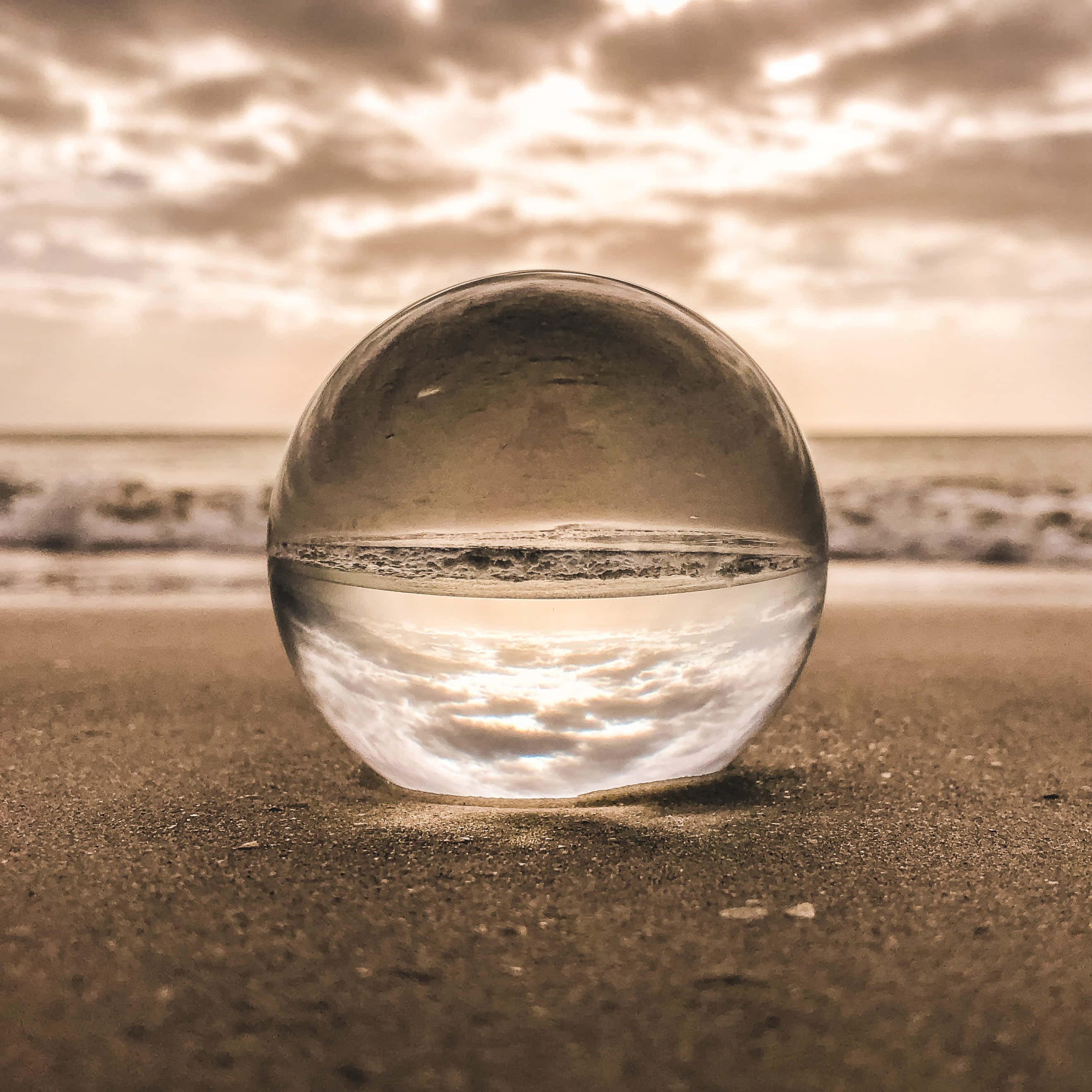 glass 桌布 壁紙 — 一個透明玻璃球放在沙灘上，反射金色日落海浪和多雲的天空。