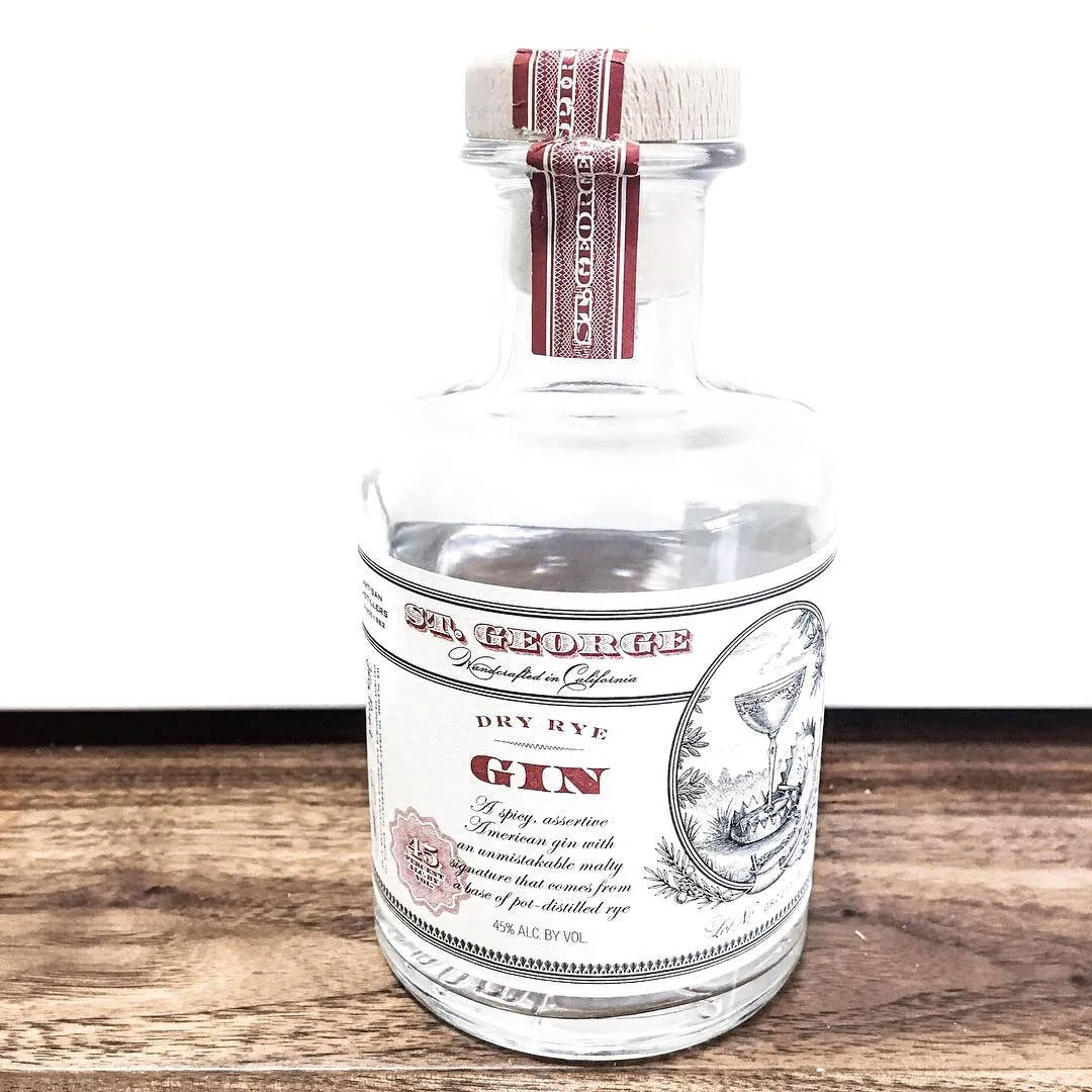 bottle 桌布 壁紙 — 一個透明玻璃瓶的 St George Dry Rye Gin，配有木蓋和復古標籤，放在白色背景下的質樸木質表面上。
