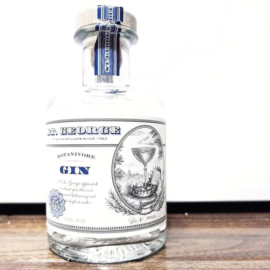 bottle 桌布 壁紙 — 一個透明玻璃瓶的 St George Botanivore Gin，帶有藍白標籤，放在木質表面上，背景為純白色。