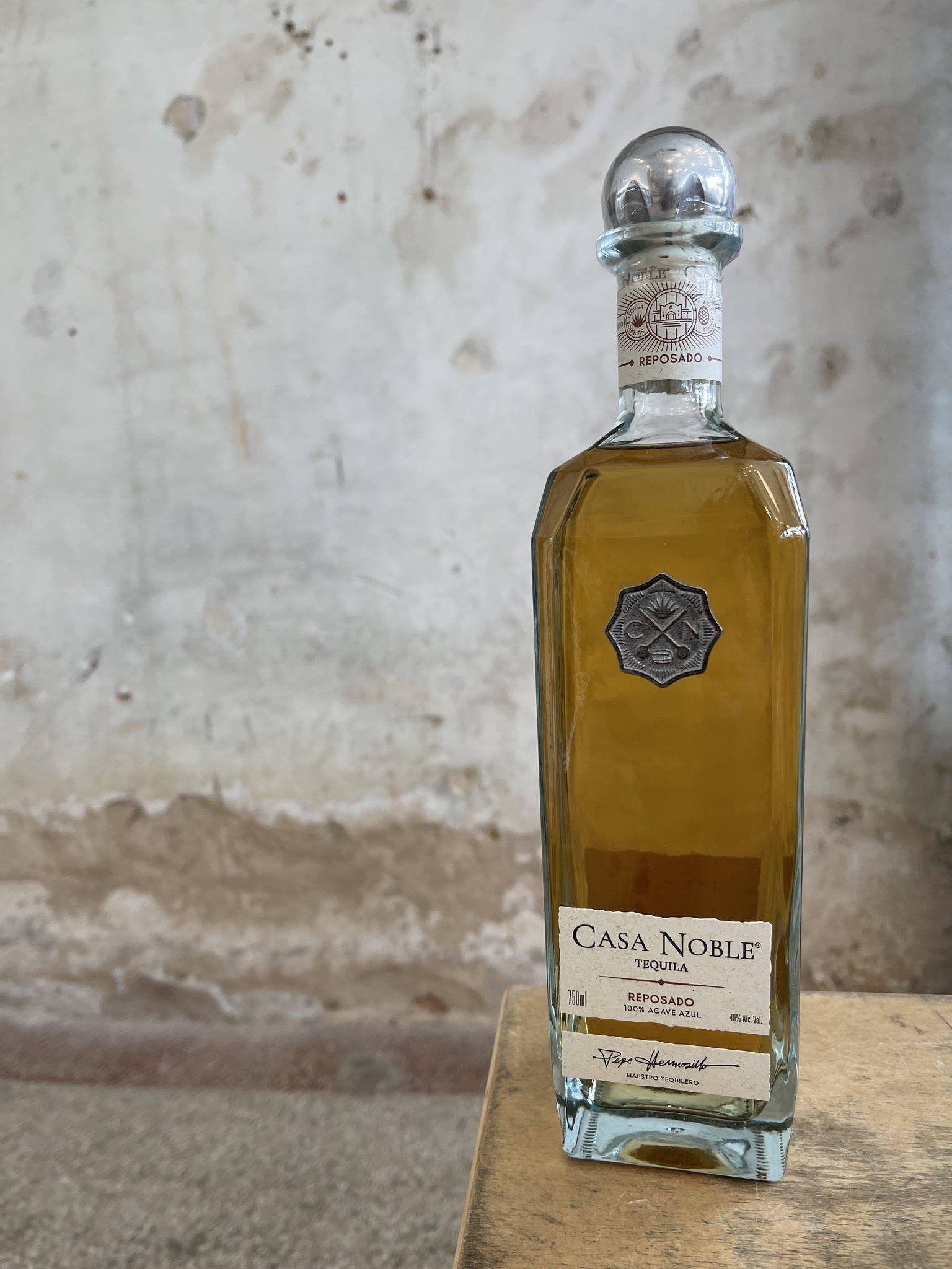 桌布 壁紙 — 一個透明玻璃瓶的Casa Noble Reposado龍舌蘭酒，裝有琥珀色液體，放在木質表面上，背景是風化的混凝土牆。