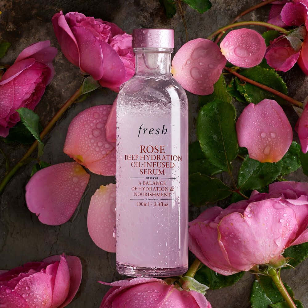 Rose Oil 桌布 壁紙 — 一個透明玻璃瓶的Fresh Rose Deep Hydration Serum，被鮮豔的粉紅玫瑰和帶露水的綠色葉子包圍，放在有紋理的石頭表面上。