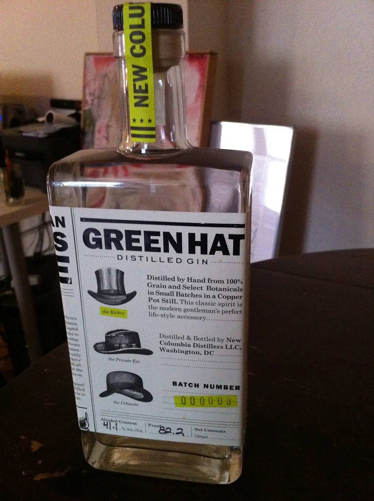 Gin 桌布 壁紙 — 一個透明玻璃瓶的Green Hat Distilled Gin，帶有黃色「New Columbia」頸標，放在昏暗房間的深色桌子上。