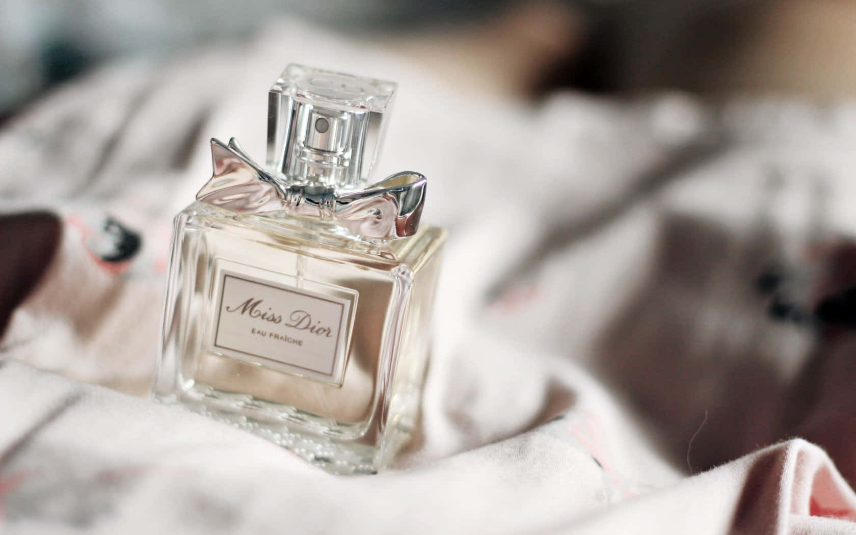 Dior 動態 桌布 壁紙 — 一個透明玻璃的 Miss Dior 香水瓶，帶有銀色蝴蝶結，優雅地放置在柔軟的皺褶布料上。