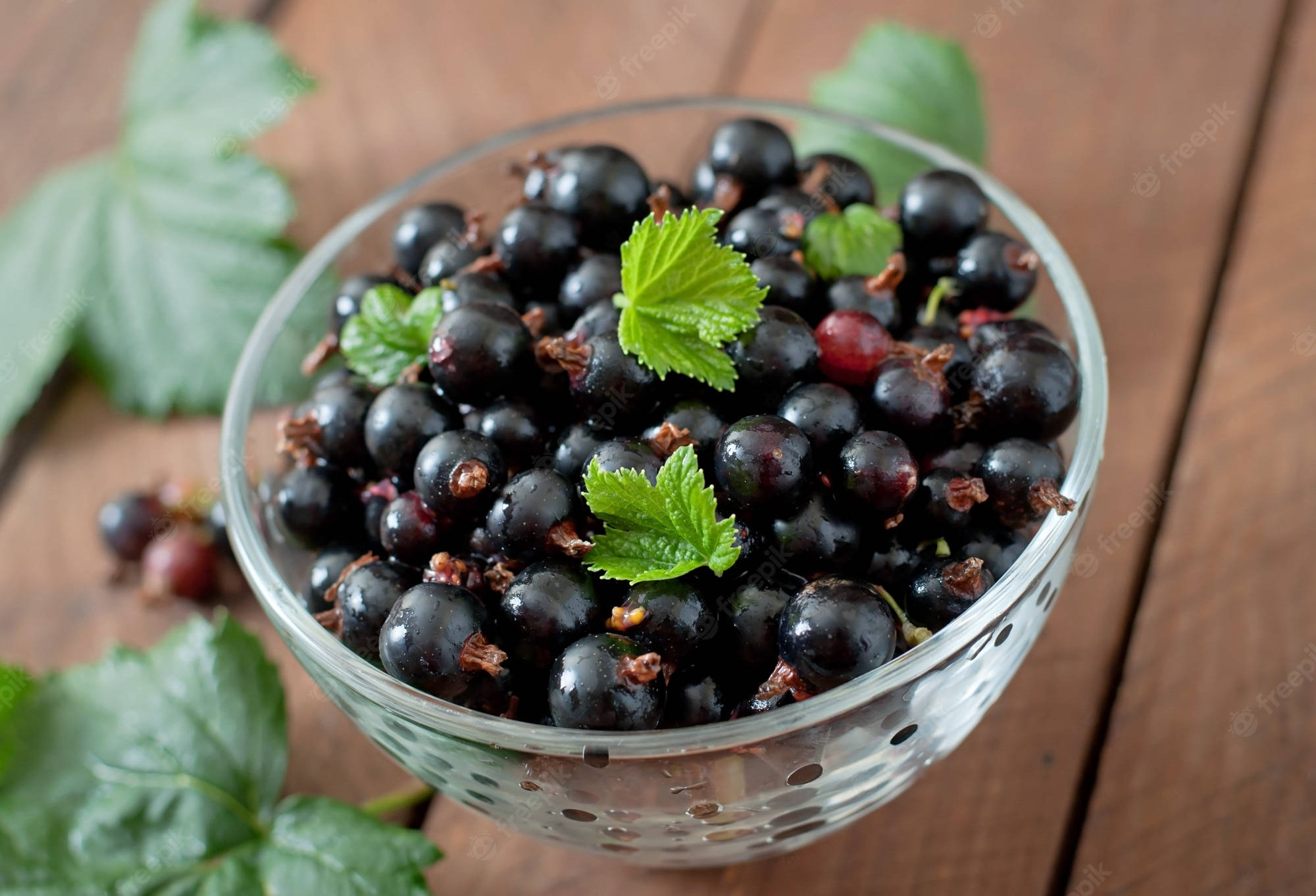 Berry 桌布 壁紙 — 一個透明玻璃碗盛滿光澤的黑加侖子和新鮮綠葉，放在質樸的木桌上。
