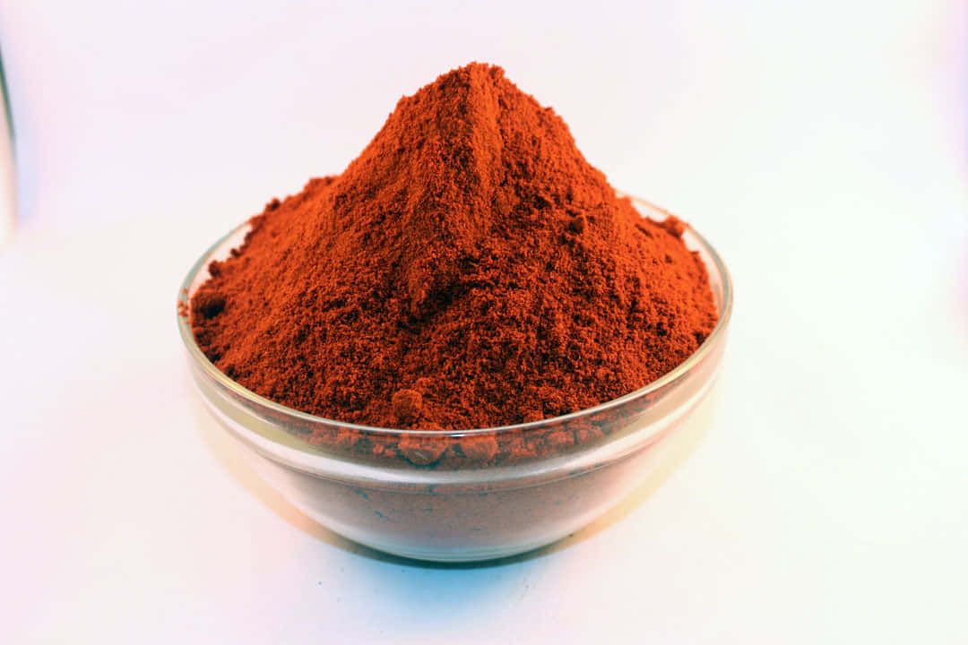 Red Chili Powder 桌布 壁紙 — 一個透明玻璃碗盛裝著一堆鮮豔的細磨紅棕色香料，背景是明亮的白色。