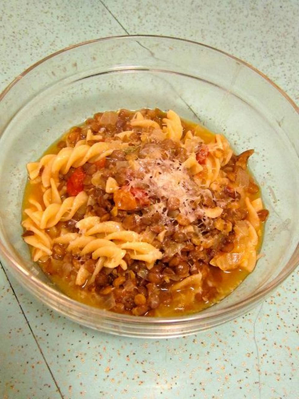 Healthy Food 桌布 壁紙 — 一個透明玻璃碗盛裝螺旋義大利麵，搭配豐富的扁豆番茄醬，上面撒上磨碎的起司，放在有斑點的檯面上。