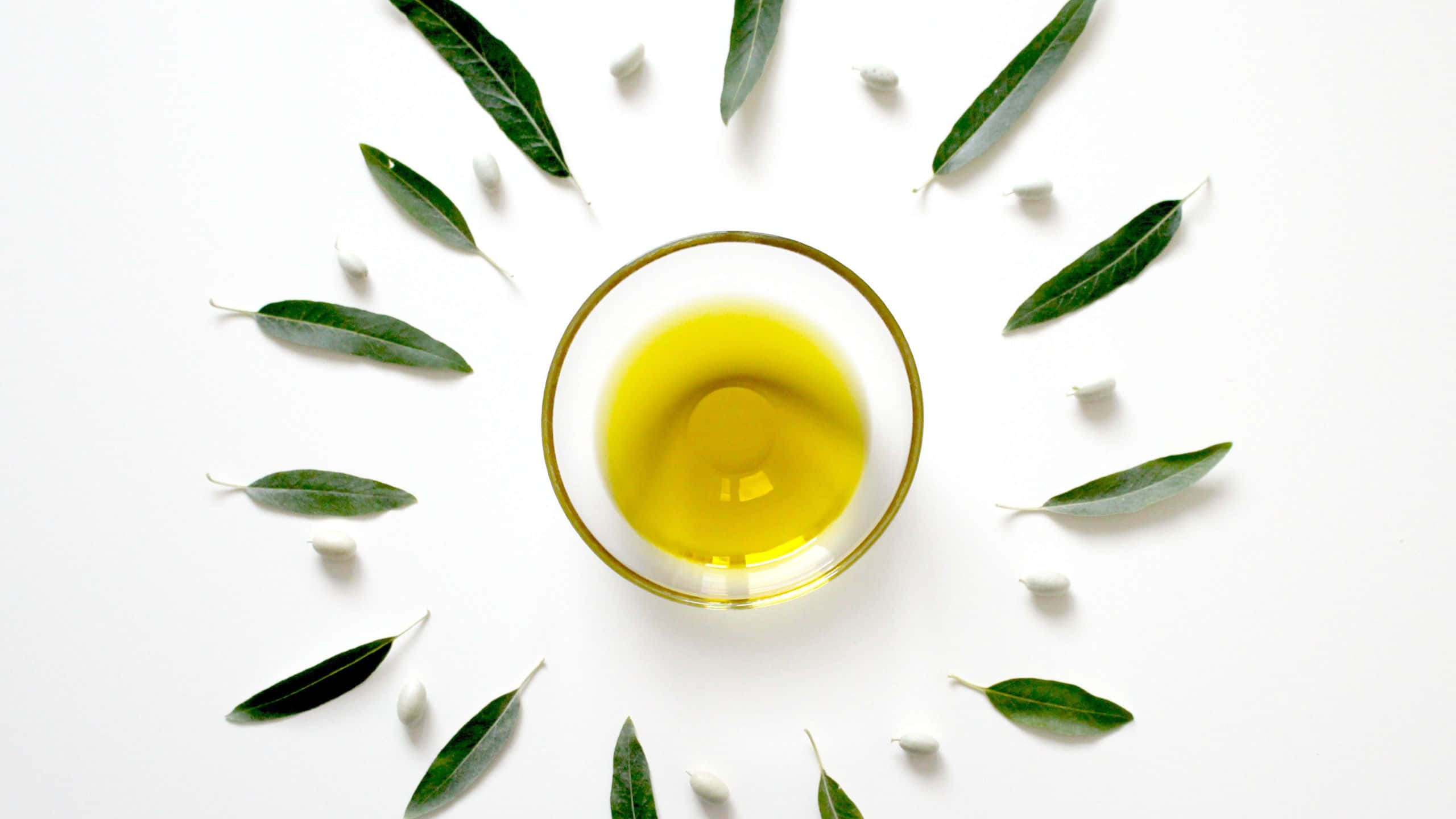 Oil 桌布 壁紙 — 一個透明玻璃碗盛載著金黃色的橄欖油，周圍環繞著新鮮的綠色葉子和白色橄欖，放在明亮的白色表面上。