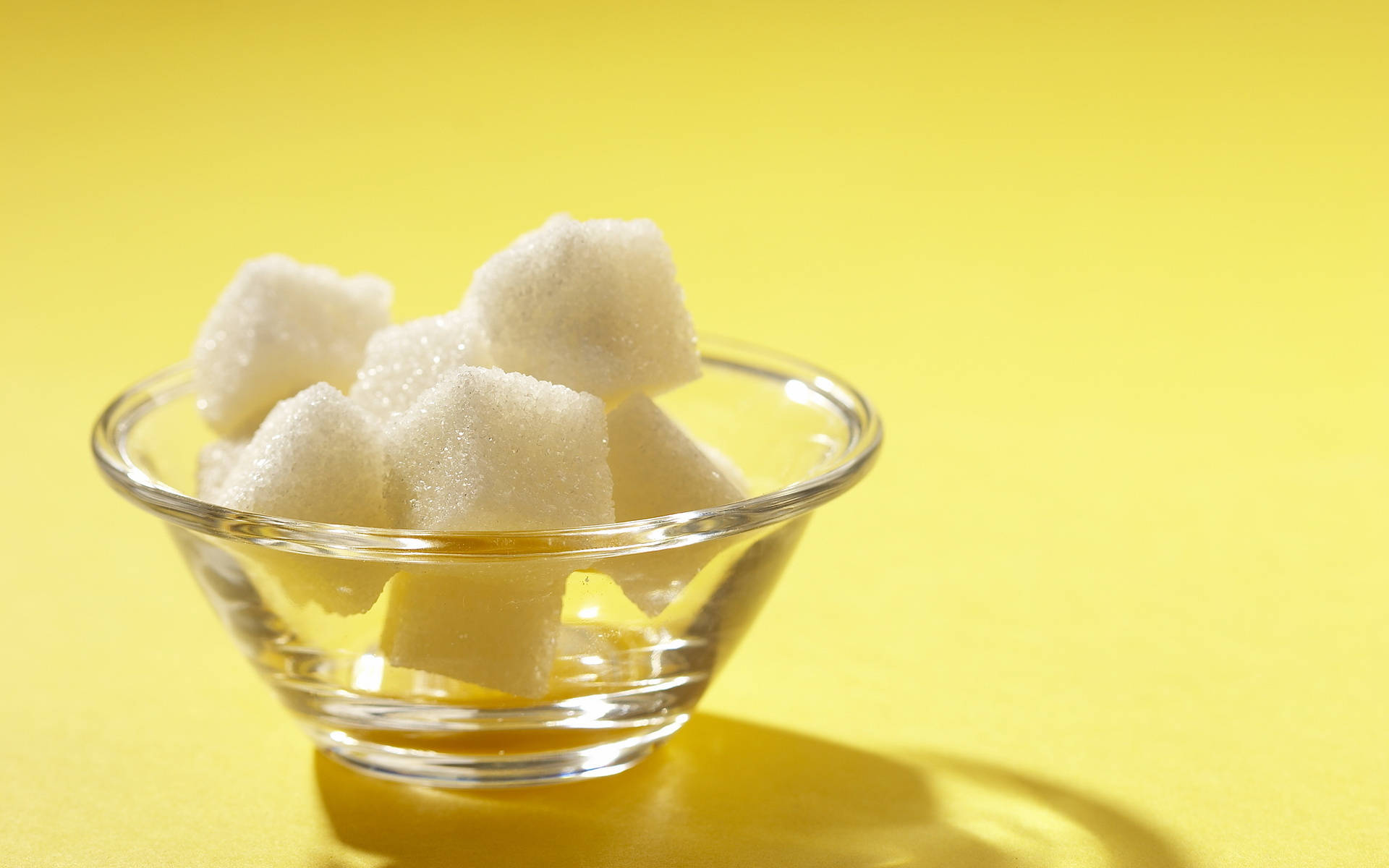 Sugar 桌布 壁紙 — 一個透明玻璃碗裝滿白色方糖，放在鮮黃色背景上，帶有柔和陰影。