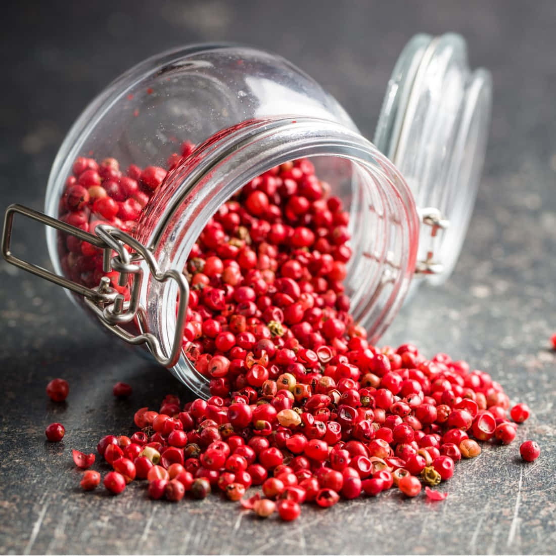 Pink Peppercorn 桌布 壁紙 — 一個透明玻璃罐翻倒，將鮮紅色的胡椒粒灑在深色紋理表面上。
