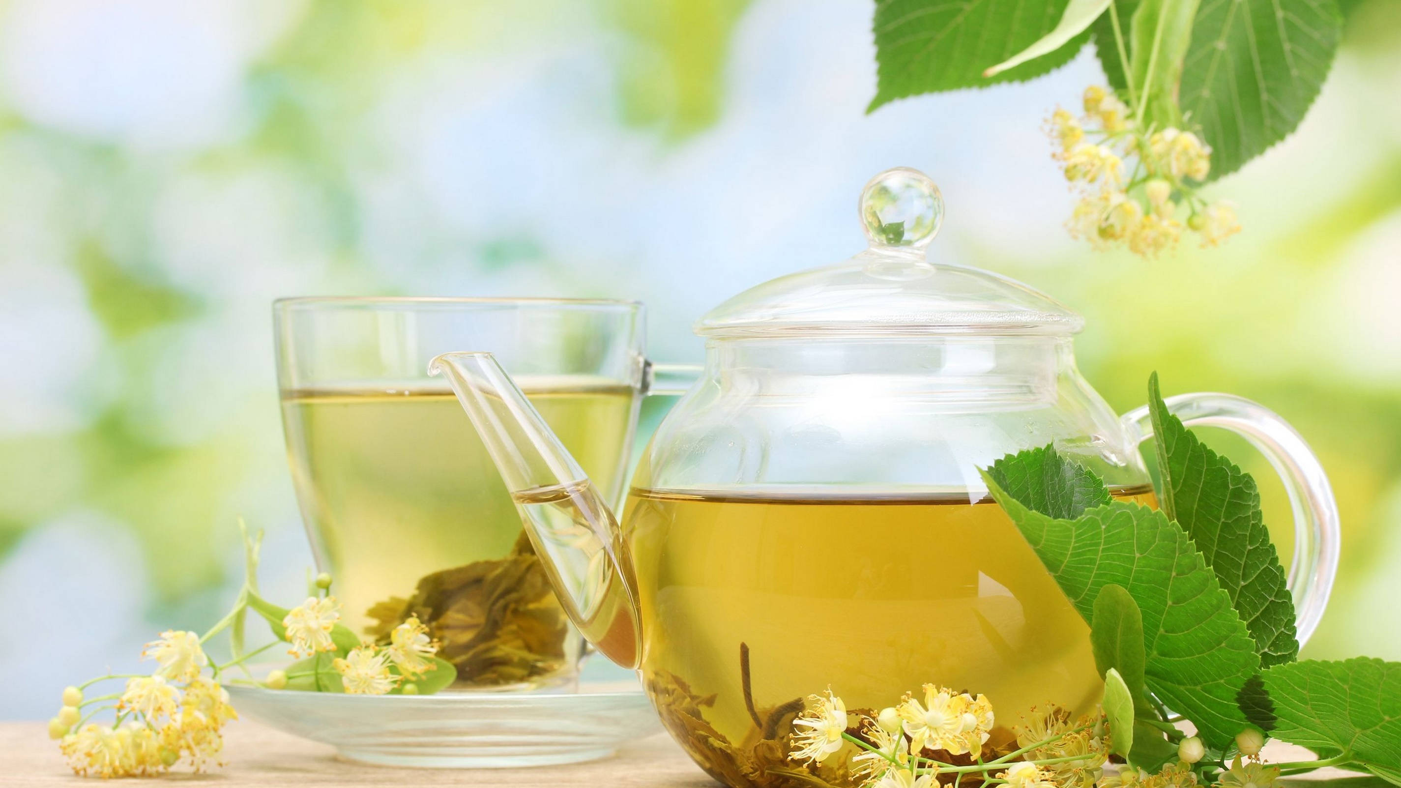 設計 桌布 壁紙 — 一個透明玻璃茶壺和杯子，盛滿金黃色的椴樹茶，周圍環繞著新鮮的綠葉和精緻的黃色花朵。