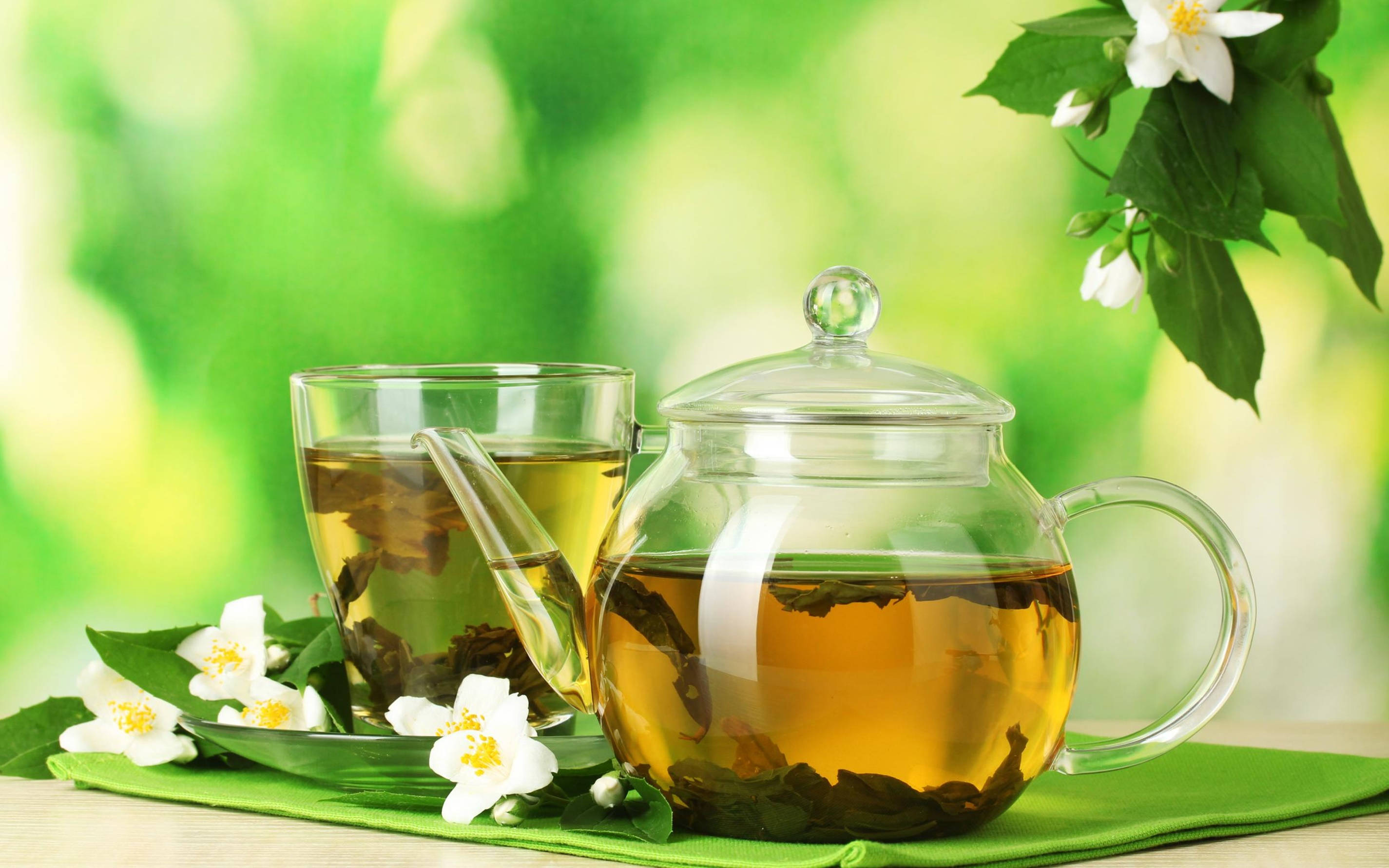 Garden 桌布 壁紙 — 一個透明玻璃茶壺和杯子盛滿金黃色茉莉花茶，放在綠色布上，旁邊有新鮮的白色花朵和葉子。