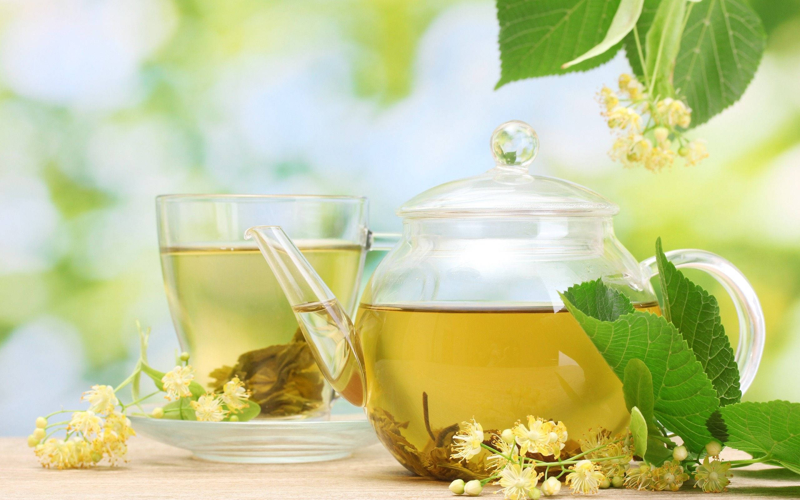 Health 桌布 壁紙 — 一個透明玻璃茶壺和杯子盛滿金黃色的椴樹茶，放在木質表面上，周圍是新鮮的綠葉和精緻的黃色椴樹花。