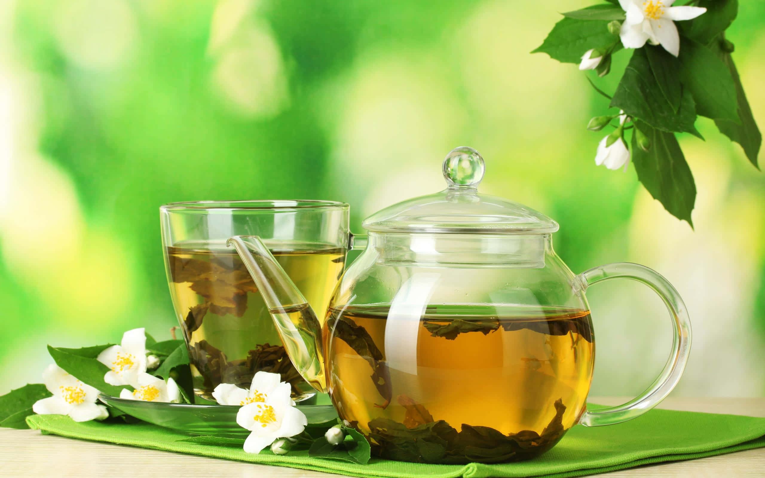 Floral Tea 桌布 壁紙 — 一個透明玻璃茶壺和杯子盛滿金黃色茉莉花茶，放在綠色布上，旁邊有新鮮的白色茉莉花和葉子，背景是柔和的綠色模糊效果。