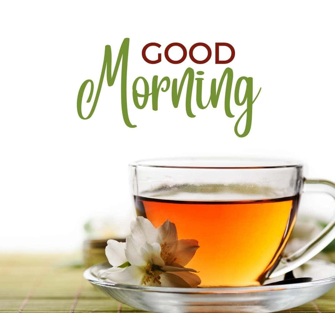 Morning Tea 桌布 壁紙 — 一個透明玻璃茶杯裝滿琥珀色茶，放在碟子上，旁邊有一朵白花，背景是綠色竹蓆，並有「GOOD Morning」文字。