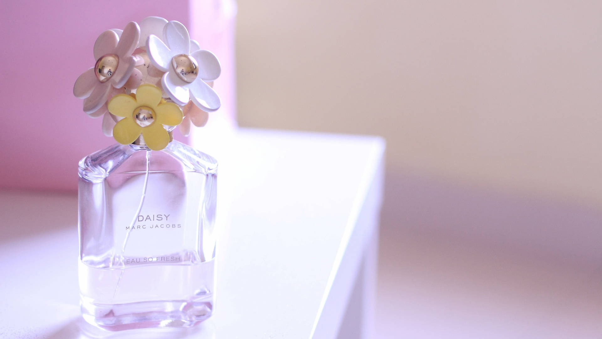 Marc Jacobs 桌布 壁紙 — 一個透明玻璃香水瓶，標有 Marc Jacobs 的 Daisy，頂部有粉彩花朵裝飾，放置在白色表面上，背景為柔和粉紅色。
