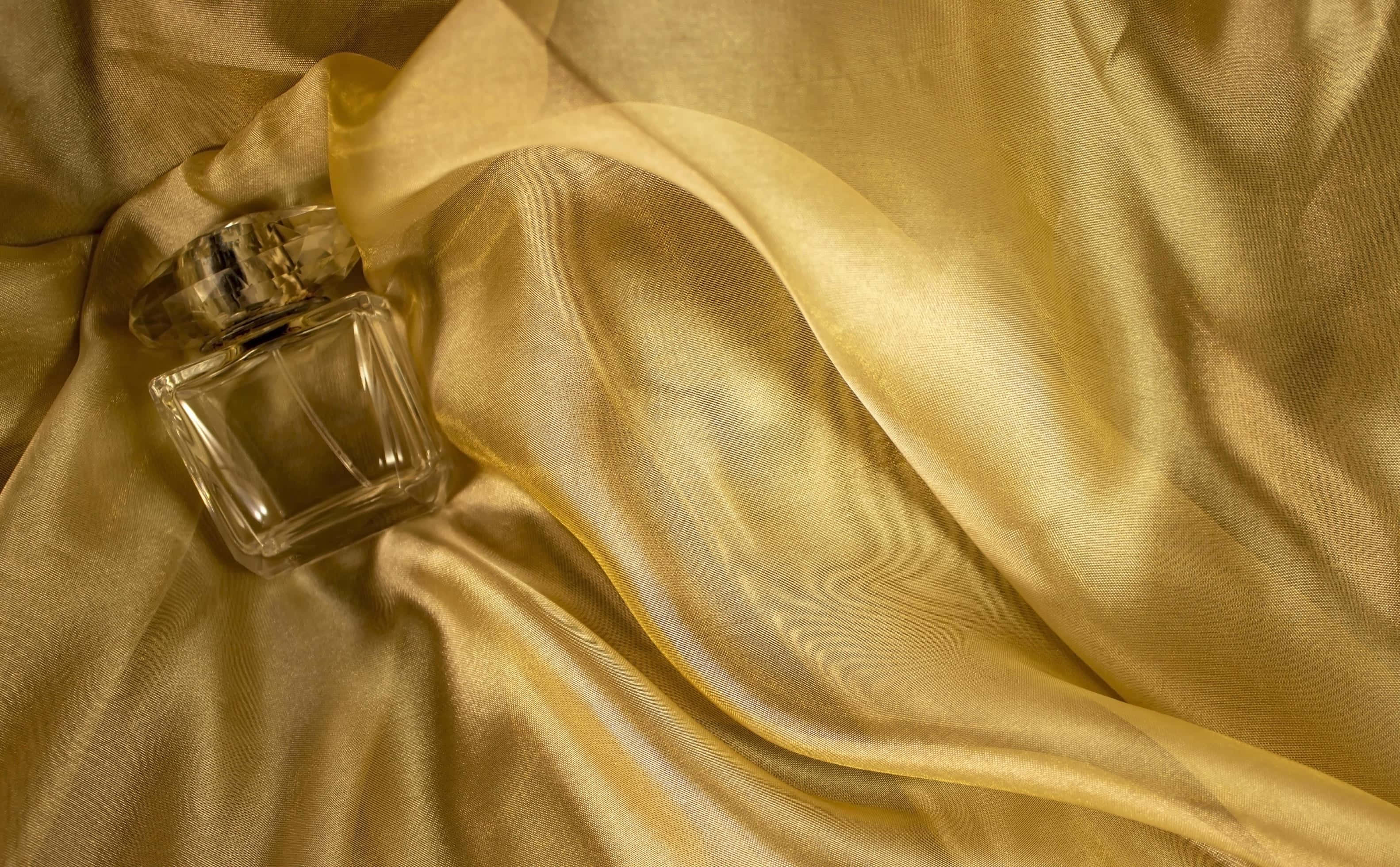 Gold Silk 桌布 壁紙 — 一個透明玻璃香水瓶放在豪華的金色絲綢布料上，帶有柔軟的褶皺和閃爍的高光。