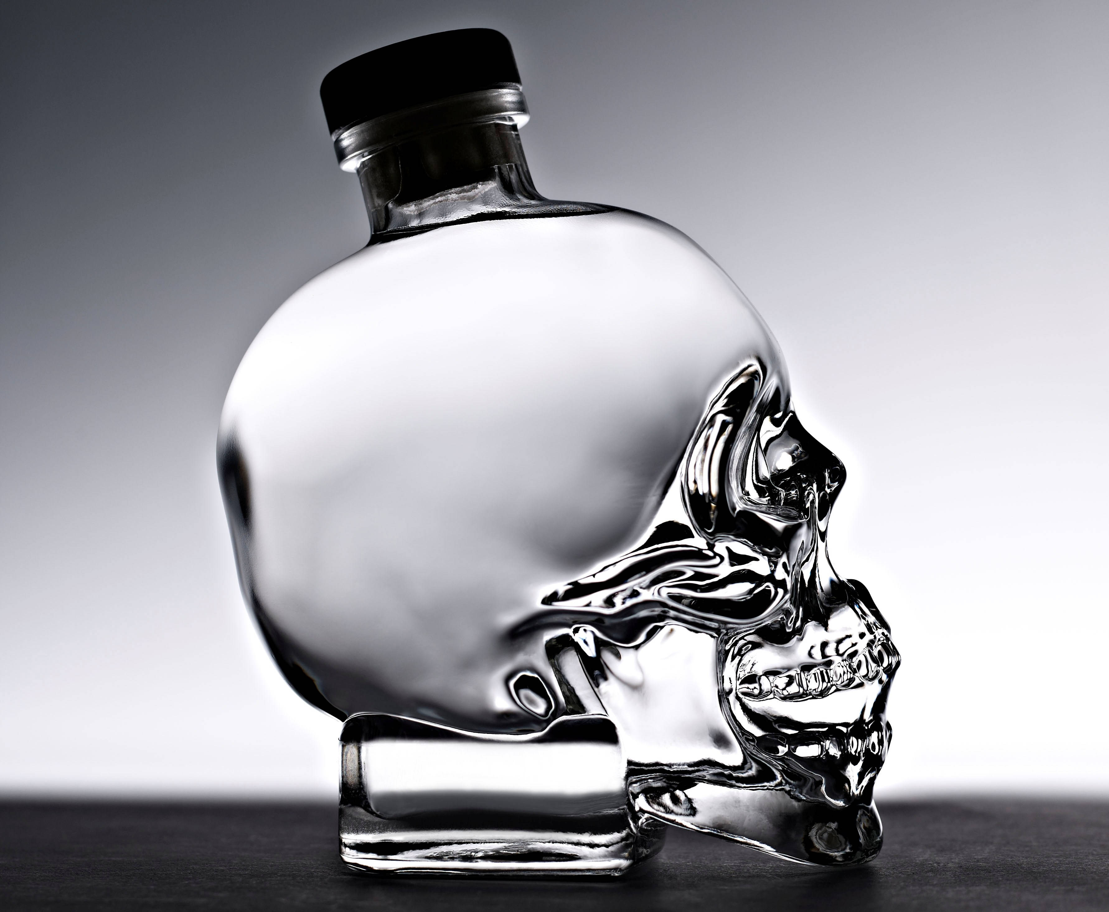 Drink 桌布 壁紙 — 一個透明玻璃骷髏形狀的瓶子，帶有黑色蓋子，放在深色表面上，背景為漸變灰色。