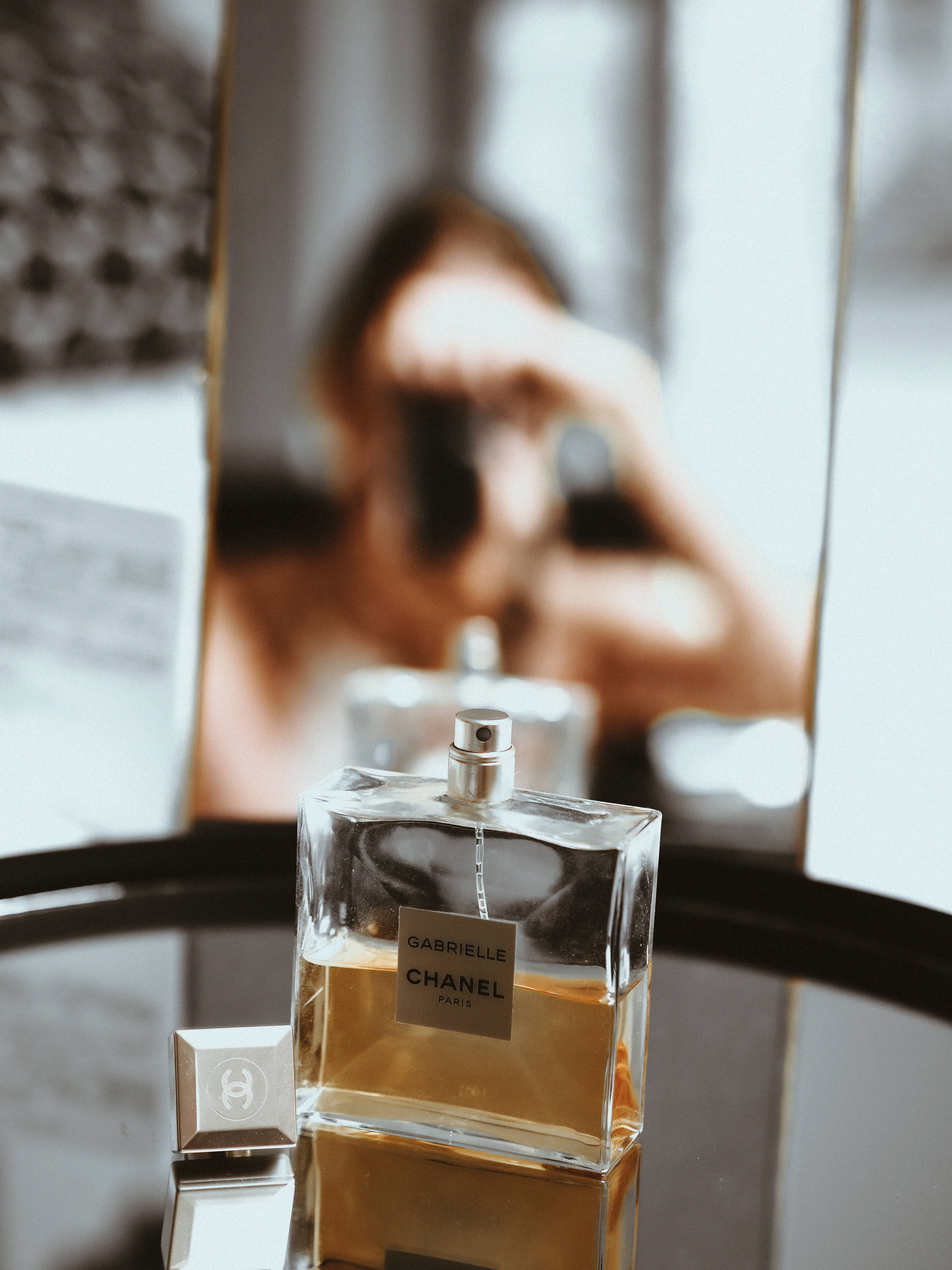 Chanel 桌布 壁紙 — 一個透明的 Chanel Gabrielle 香水瓶放在反光表面上，背景中有一個模糊的人影拿著相機。