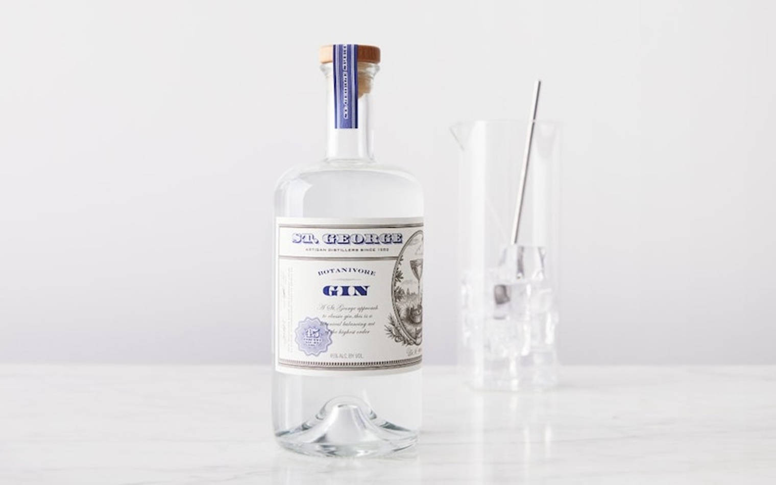 bottle 桌布 壁紙 — 一個透明的 St George Botanivore Gin 酒瓶放在白色大理石表面上，旁邊有一個玻璃雞尾酒調酒器和金屬攪拌棒。