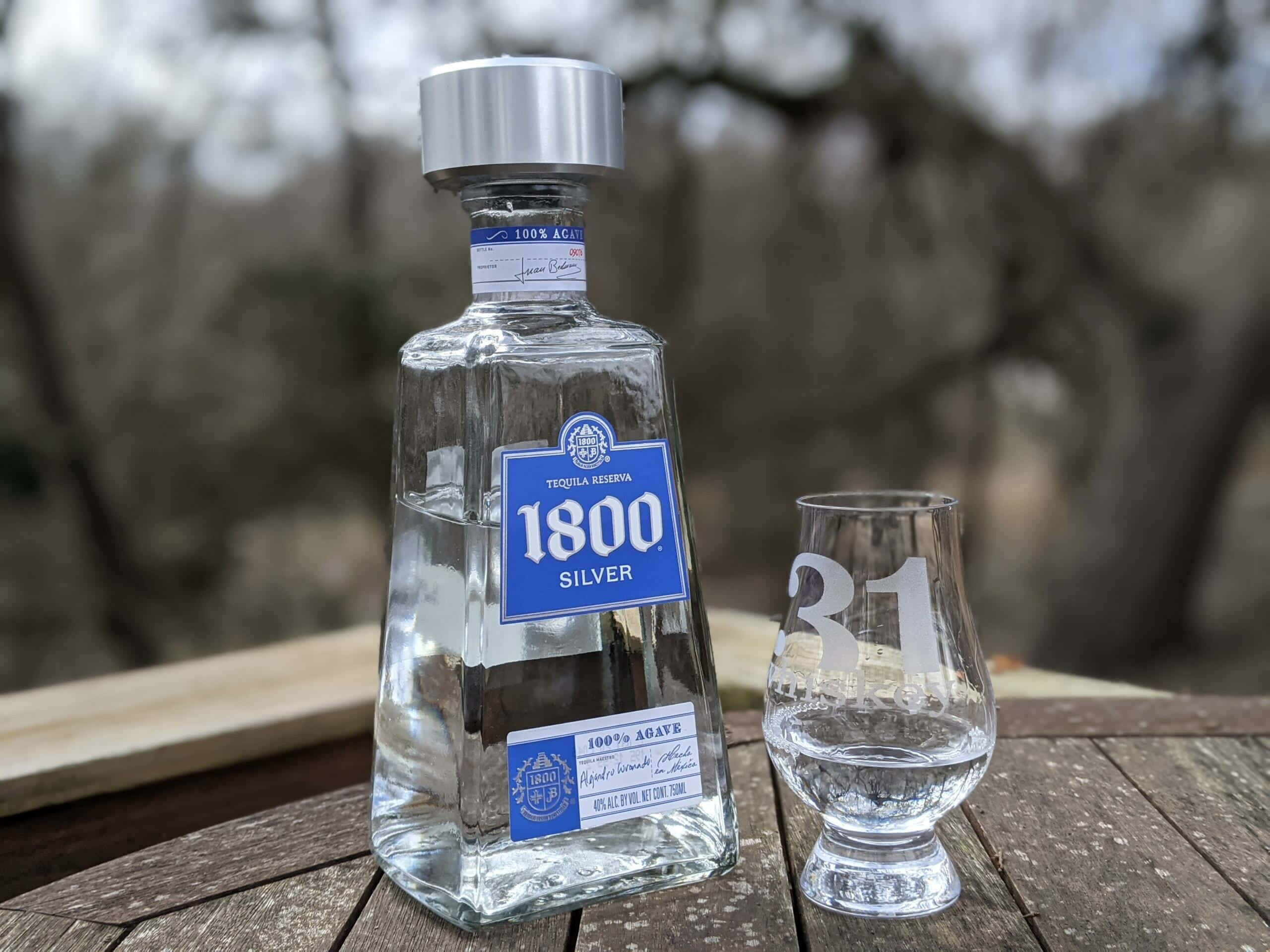 alcohol 桌布 壁紙 — 一個透明的1800 Silver龍舌蘭酒瓶和一個刻有「31」的玻璃杯放在戶外一張風化的木桌上。