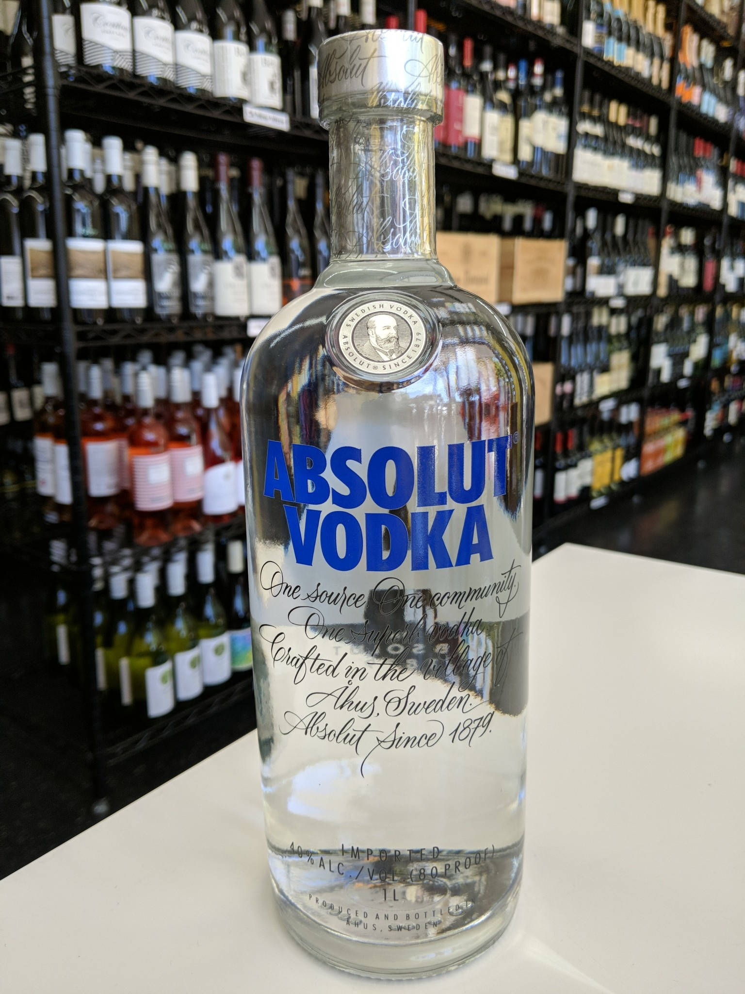 bottle 桌布 壁紙 — 一個透明的Absolut Vodka酒瓶放在白色表面上，背景中模糊的酒瓶架隱約可見。