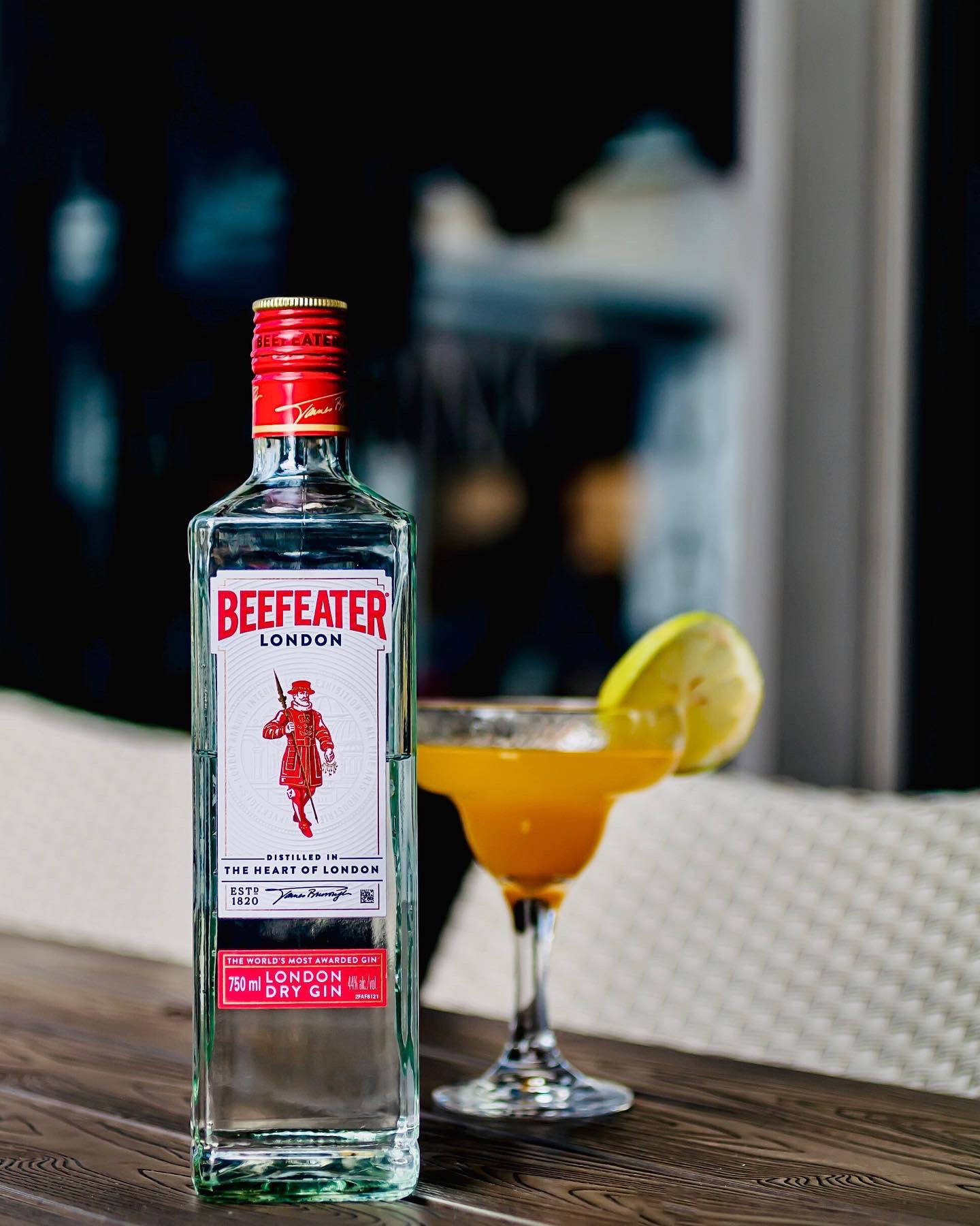 cocktail 桌布 壁紙 — 一個透明的Beefeater London Dry Gin酒瓶旁邊放著一杯充滿活力的橙色雞尾酒，上面用青檸角裝飾，放在木桌上。