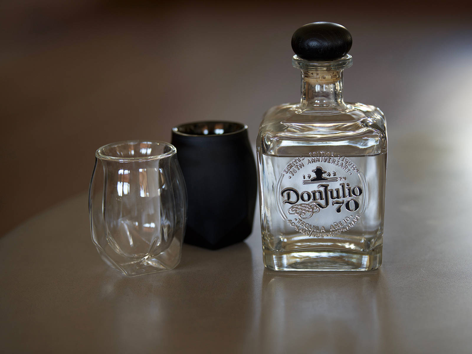 glass 桌布 壁紙 — 一個透明的Don Julio 70龍舌蘭酒瓶，黑色瓶蓋，旁邊有兩個酒杯，放在反光的木桌上。