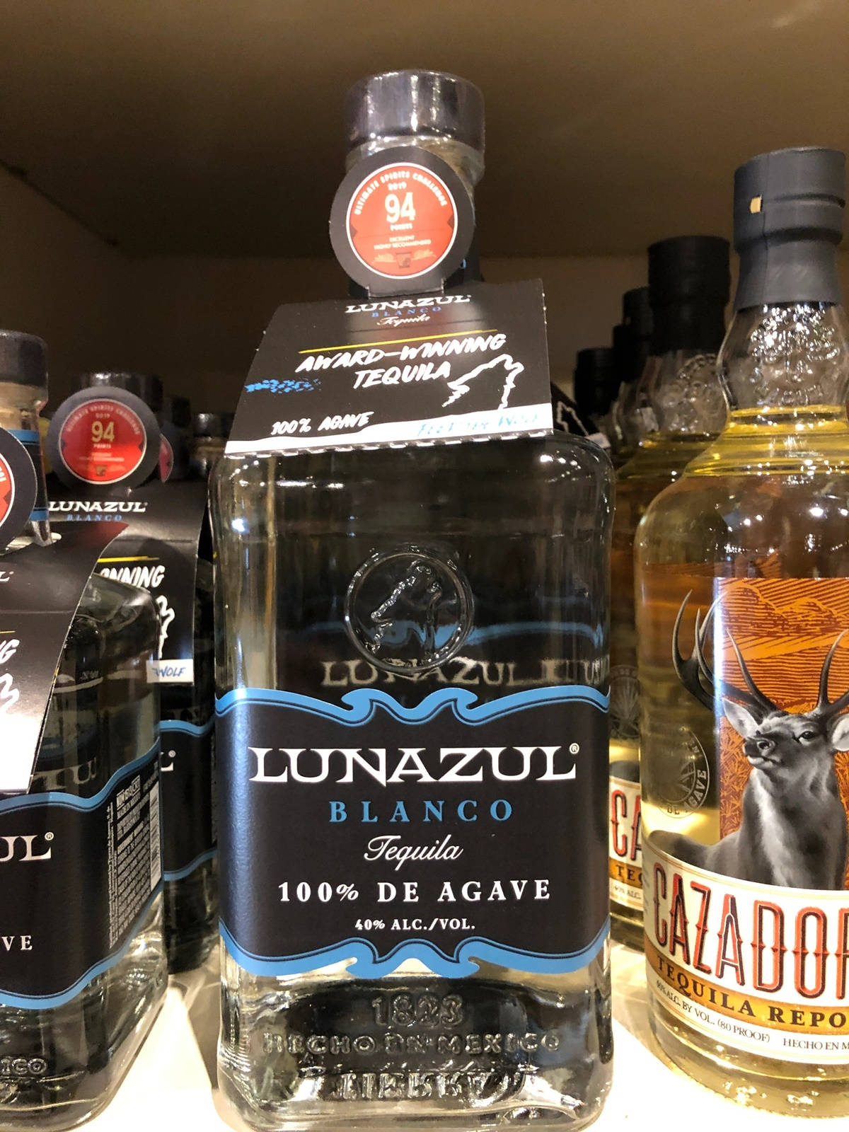alcohol 桌布 壁紙 — 一個透明的Lunazul Blanco龍舌蘭酒瓶，黑色標籤和藍色點綴，放在架子上，旁邊有其他酒瓶。