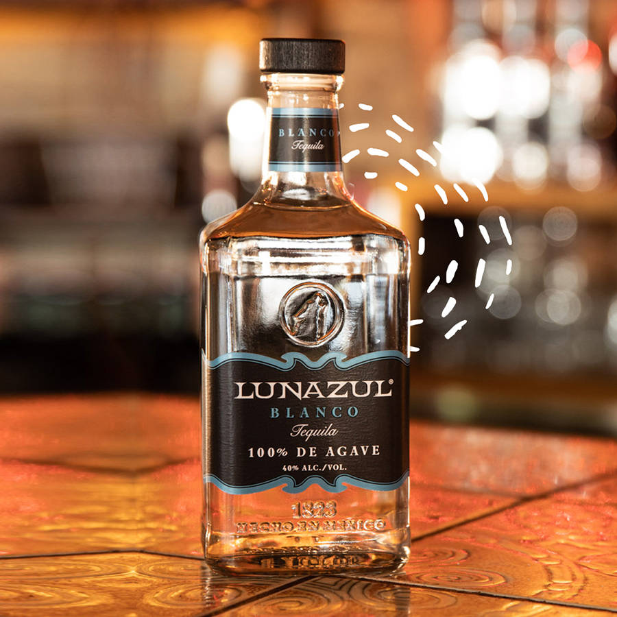 alcohol 桌布 壁紙 — 一個透明的Lunazul Blanco龍舌蘭酒瓶放在溫暖的木吧檯上，背景有模糊的玻璃器皿和金色燈光。