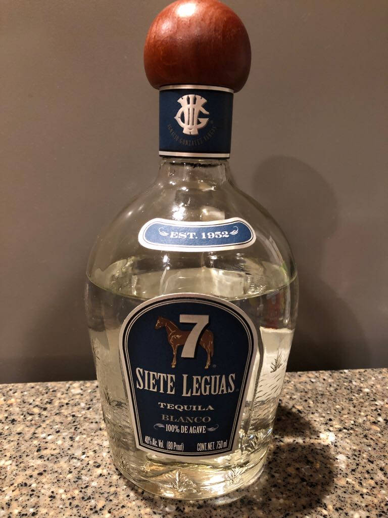 手機 桌布 壁紙 — 一個透明的Siete Leguas tequila blanco瓶子，帶有木塞，放在有斑點的檯面上，背景是素牆。