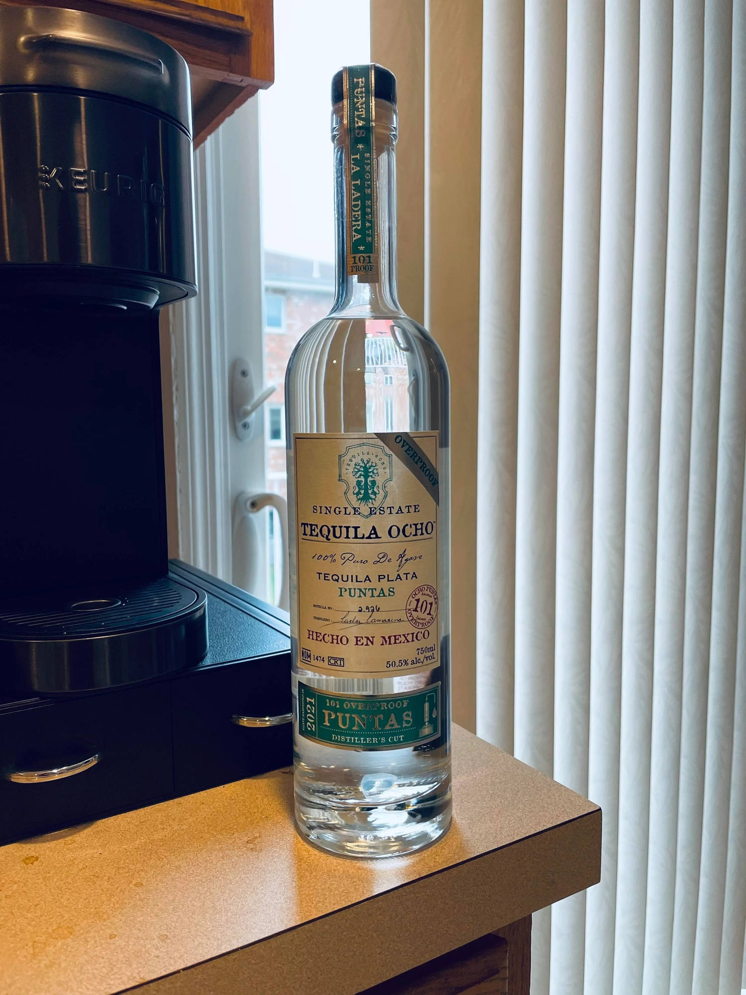 bottle 桌布 壁紙 — 一個透明的Tequila Ocho酒瓶放在木製檯面上，旁邊有一部黑色Keurig咖啡機，背景有垂直百葉窗。