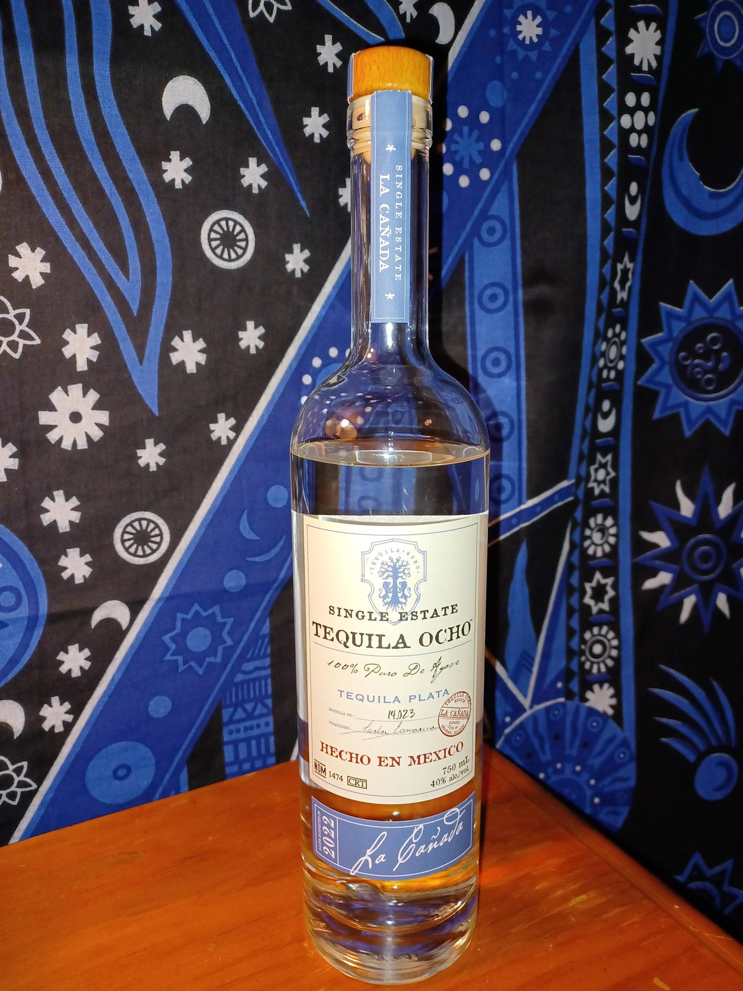 bottle 桌布 壁紙 — 一個透明的Tequila Ocho酒瓶放在木質表面上，背景是鮮豔的藍白圖案紡織品。