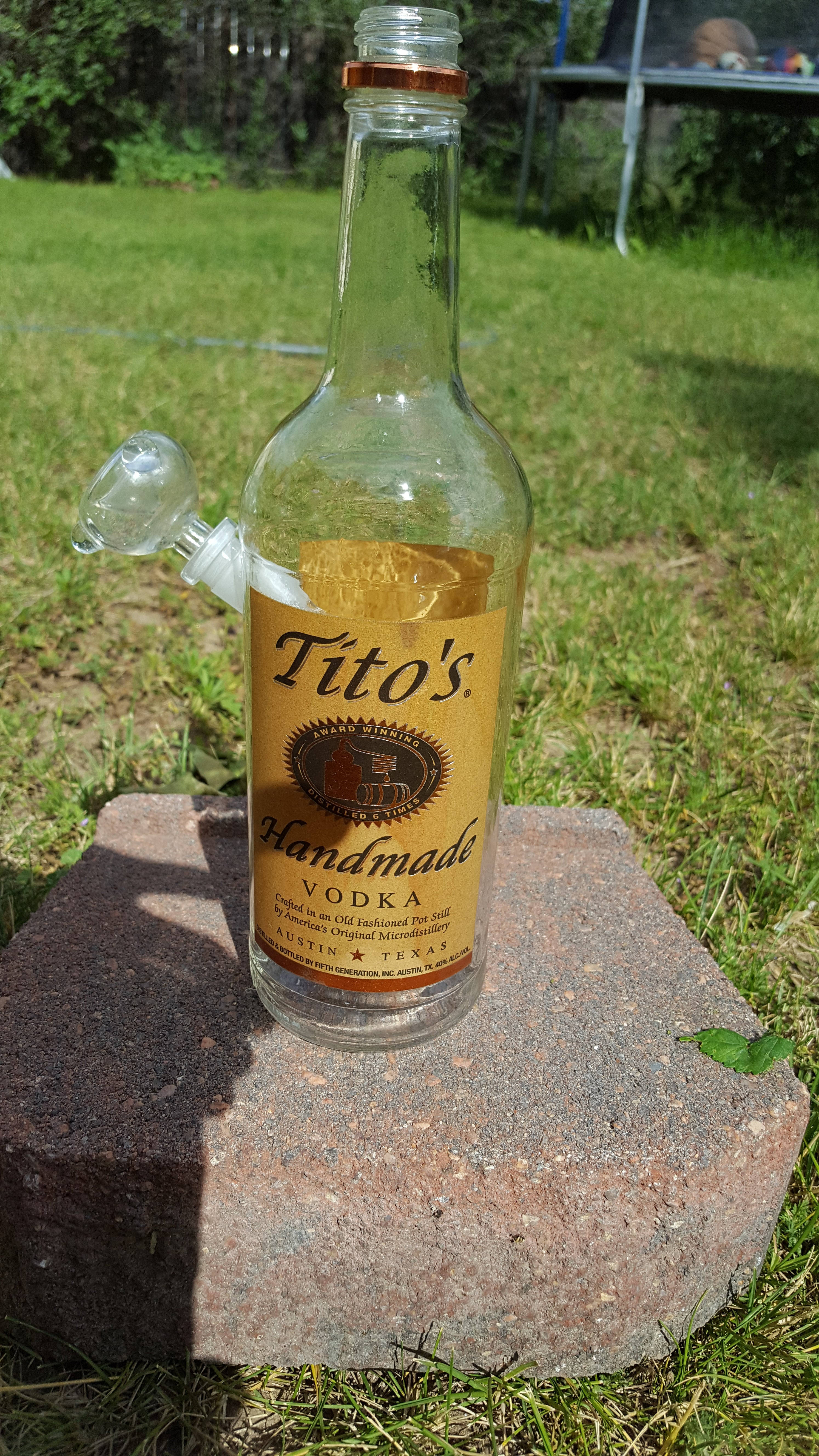 標誌 桌布 壁紙 — 一個透明的Tito's伏特加酒瓶，帶有玻璃水煙槍配件，放在陽光普照的草地上的一塊紅磚上。