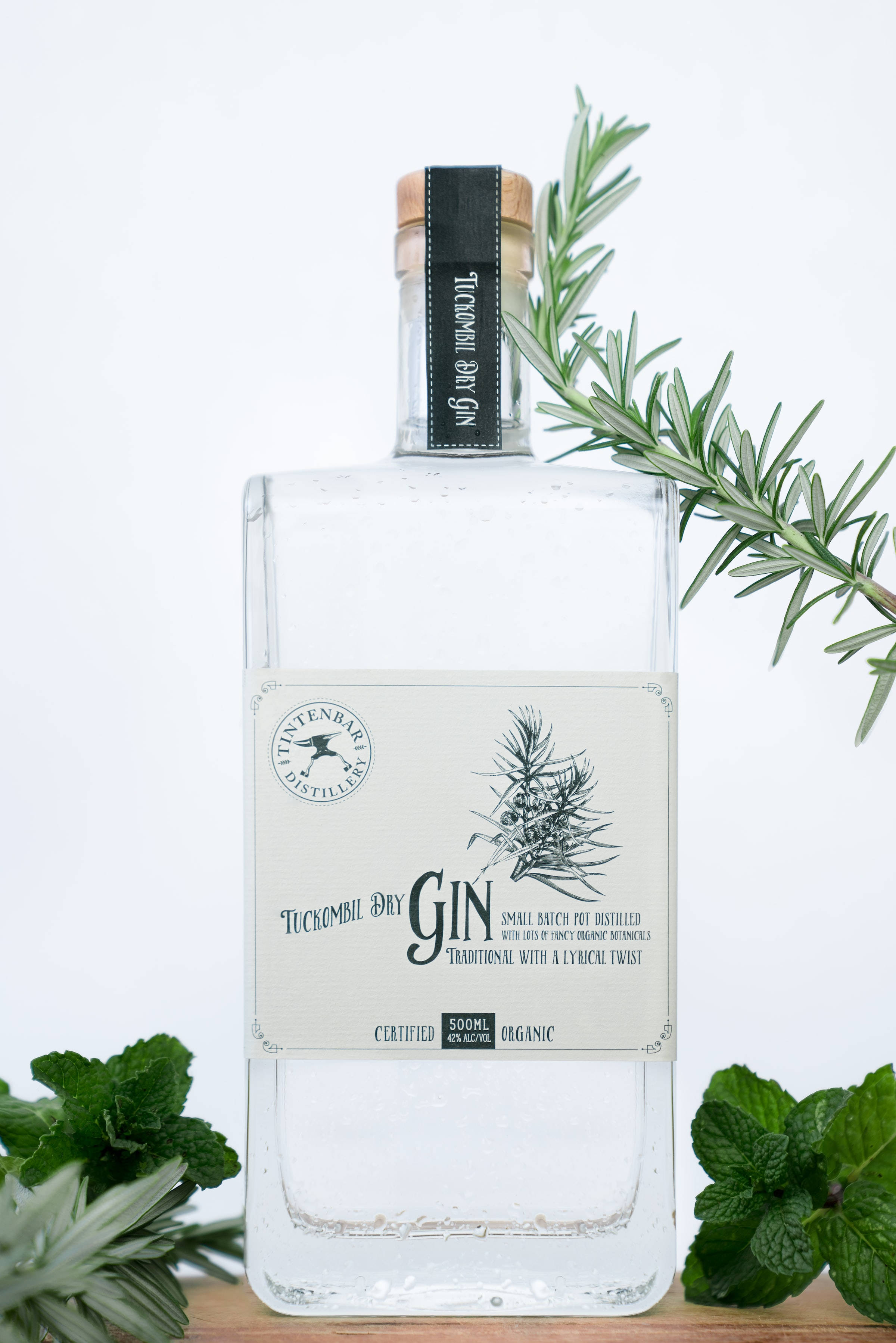 drinks 桌布 壁紙 — 一個透明的Tuckombil乾琴酒瓶放在木質表面上，周圍有新鮮迷迭香和薄荷枝，背景為白色。