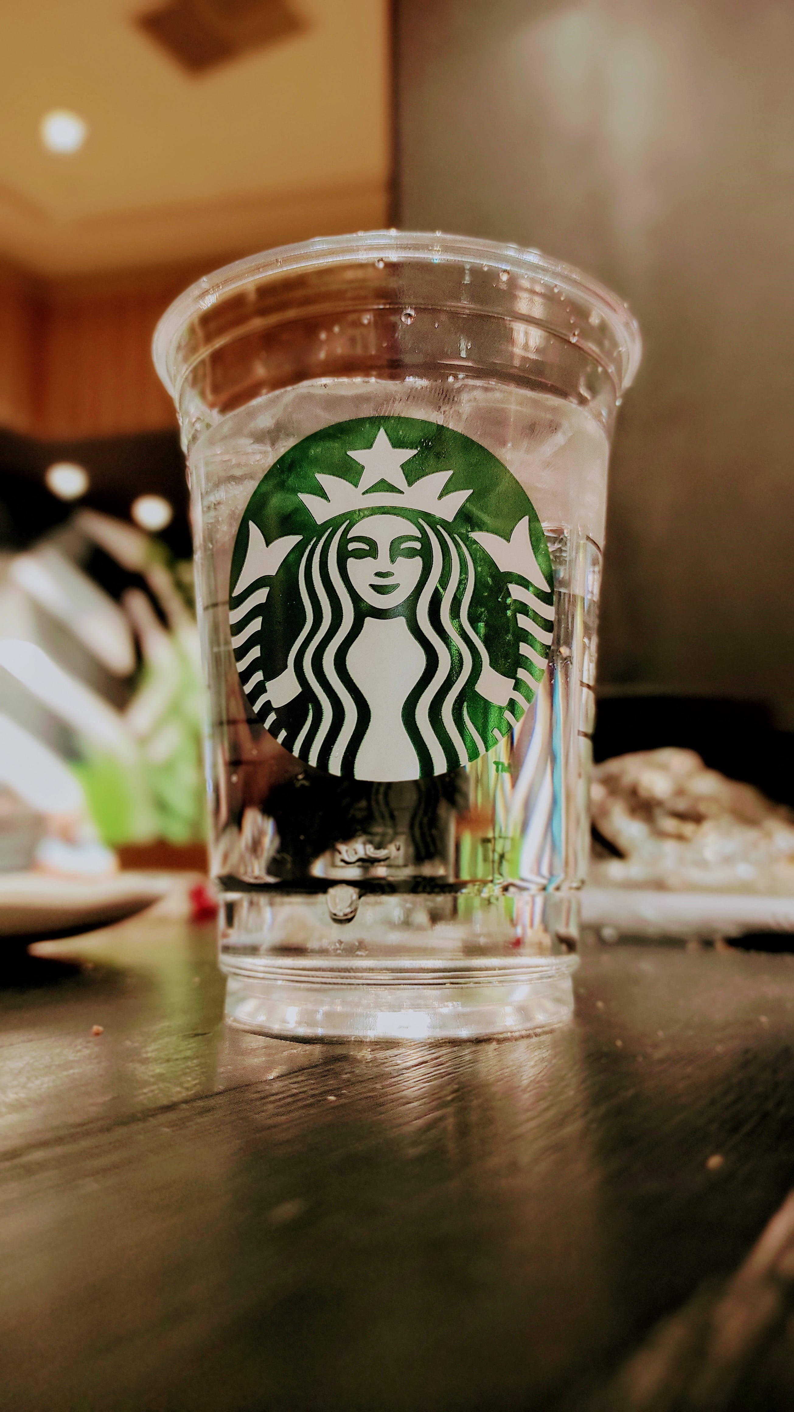 Drink Water 桌布 壁紙 — 一個透明的星巴克杯子裝滿水，放在一間燈光溫暖的咖啡店的深色木桌上。